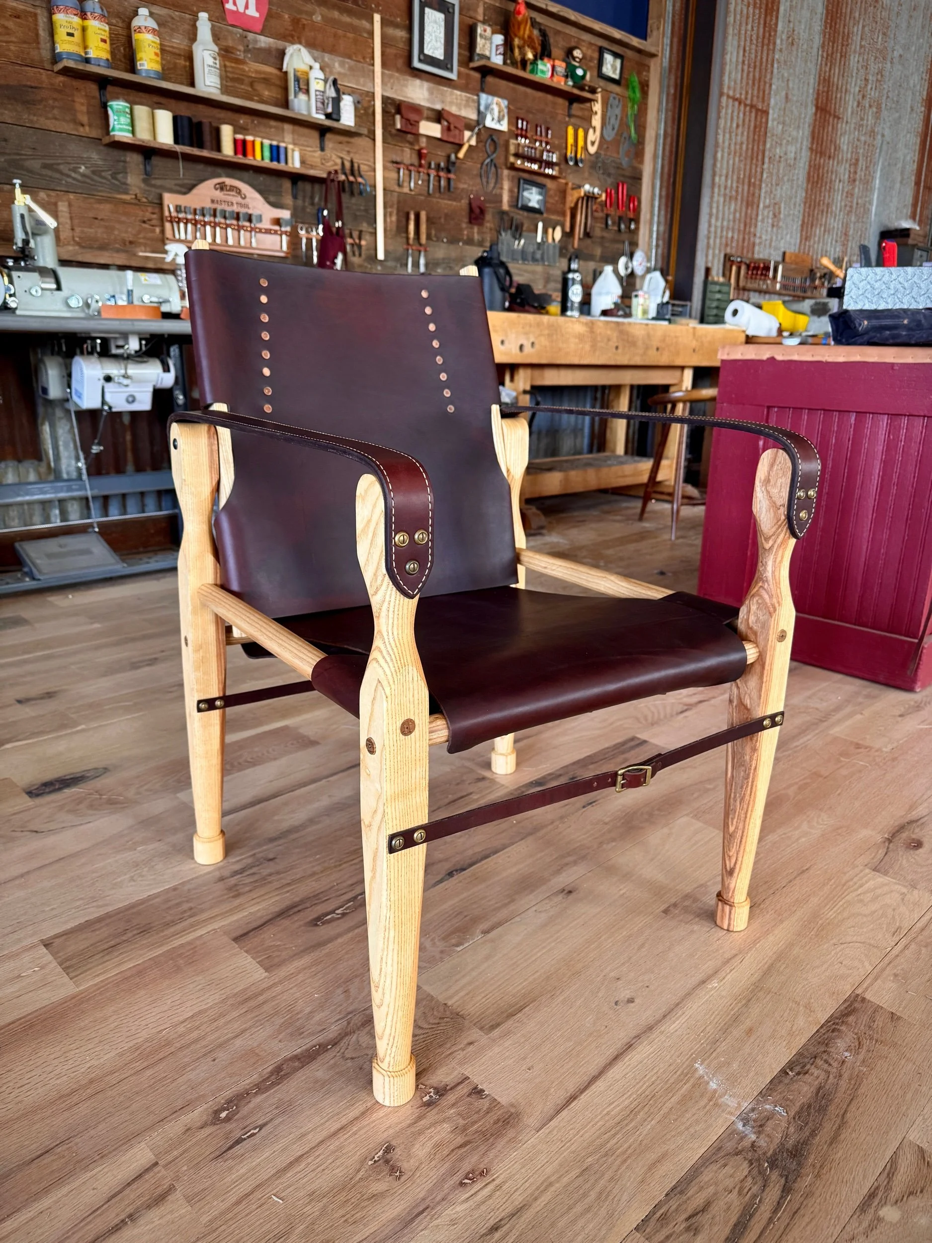 A wooden chair with leather seat and backrest, detailed with brass hardware, located in a workshop with tools and woodwork supplies in the background.