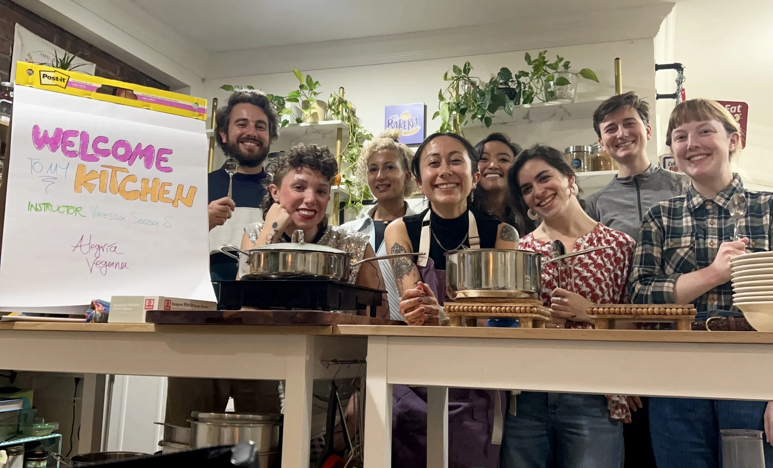 A group of people in a kitchen. All of them smiling and surrounded by kitchen supplies