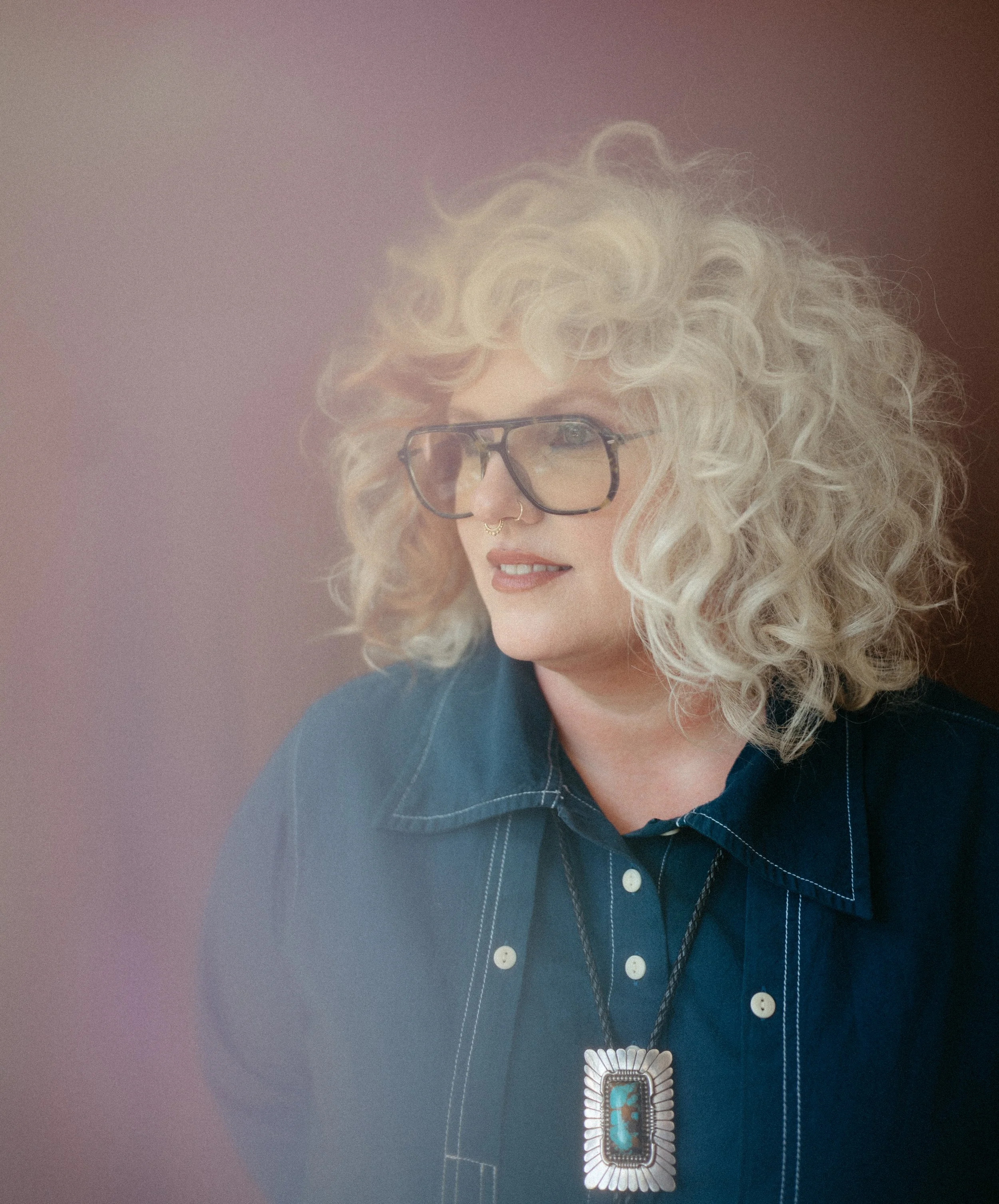  Editorial portrait of a woman with short platinum blonde curly hair, wearing a vibrant patterned jumpsuit. Styled for a salon branding shoot, showcasing voluminous curls and natural texture on a soft blue backdrop.