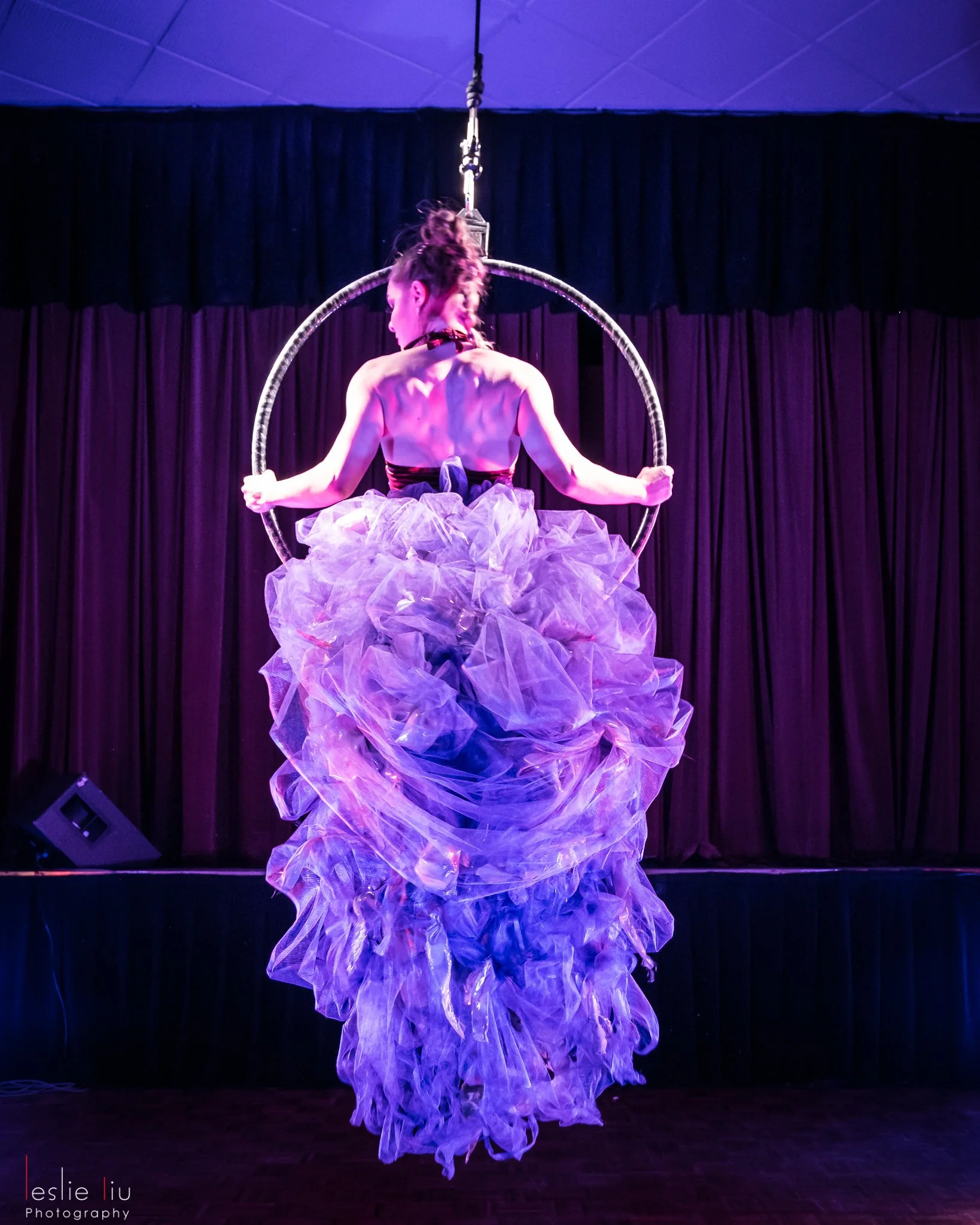 Aerial performer suspended upside-down in a glowing Lyra hoop, pouring drinks midair amid theatrical lighting and smoke.