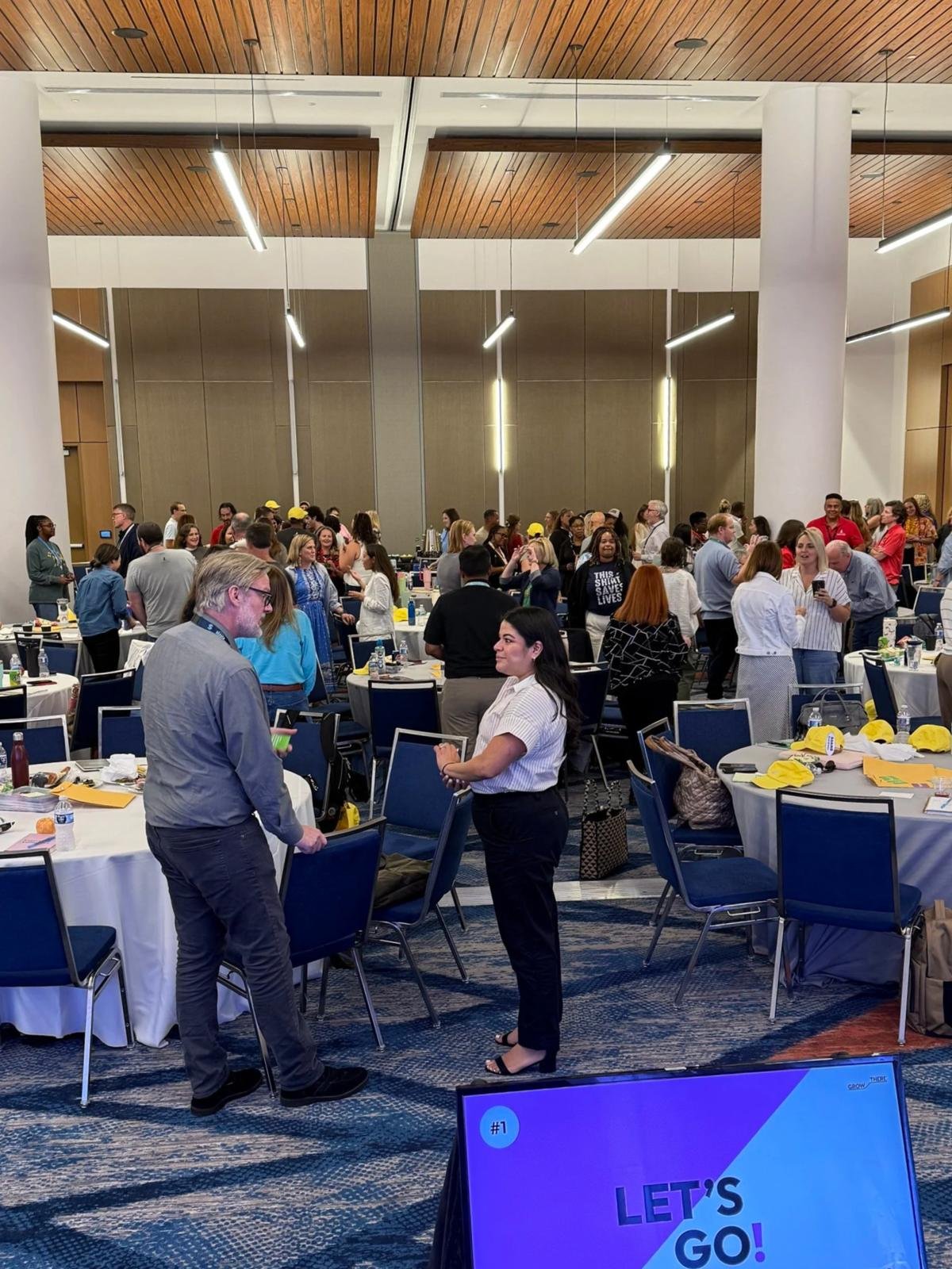 Large room of attendees standing and connecting during an interactive activity, with a Grow There “Let’s Go!” slide in front