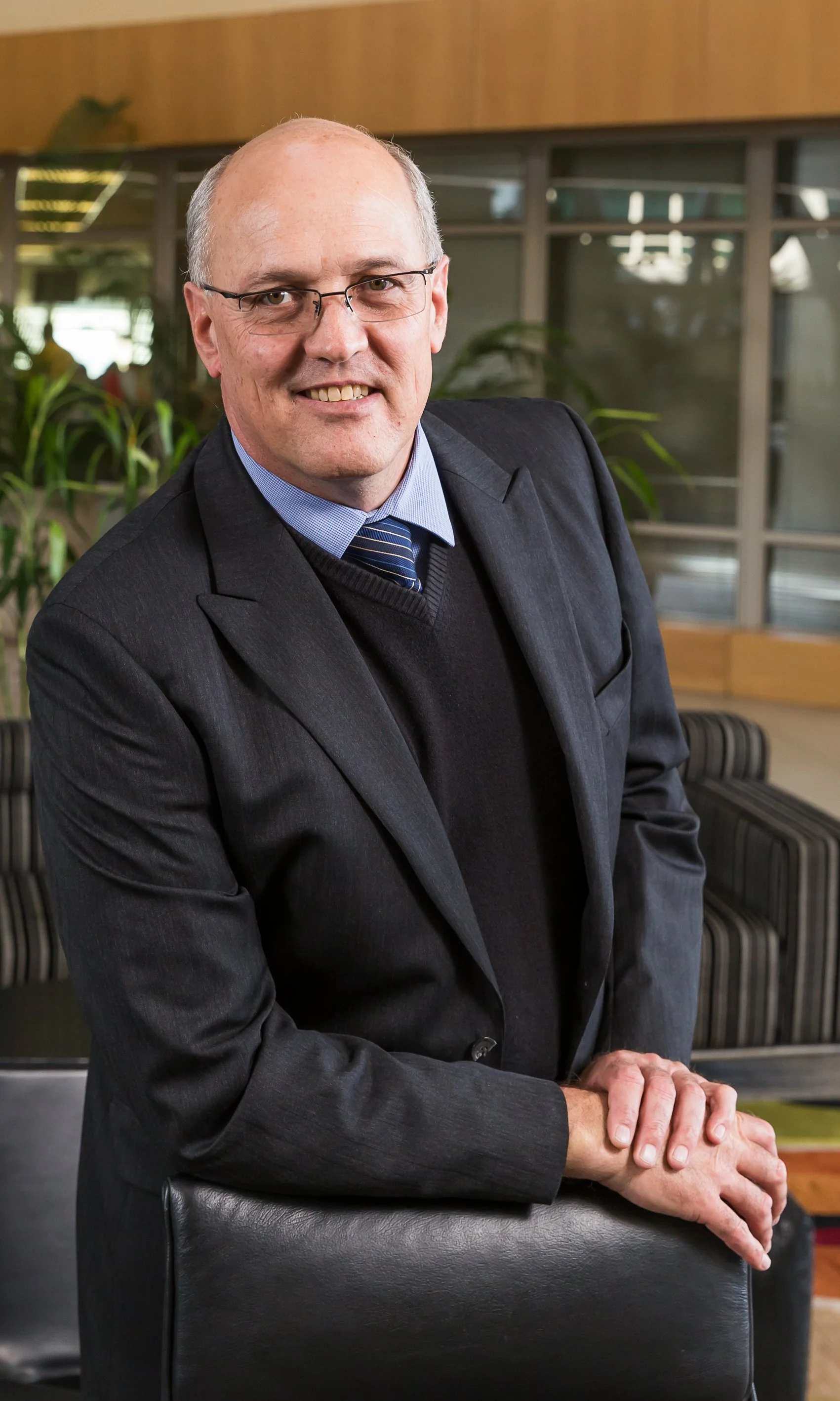A middle-aged man wearing glasses, a dark suit, a light blue shirt, and a blue tie with diagonal stripes, standing in a modern office space with large windows and plants in the background. He is smiling and resting his hands on the back of a black le