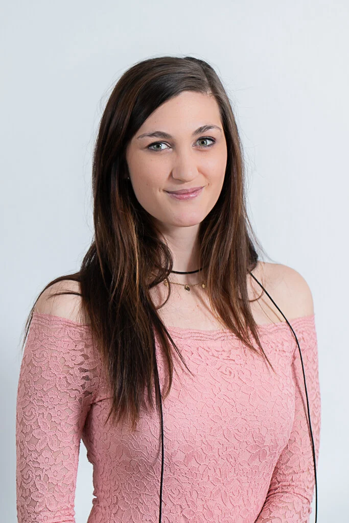 A young woman with long brown hair and fair skin wearing a pink off-the-shoulder lace top and listening to headphones.