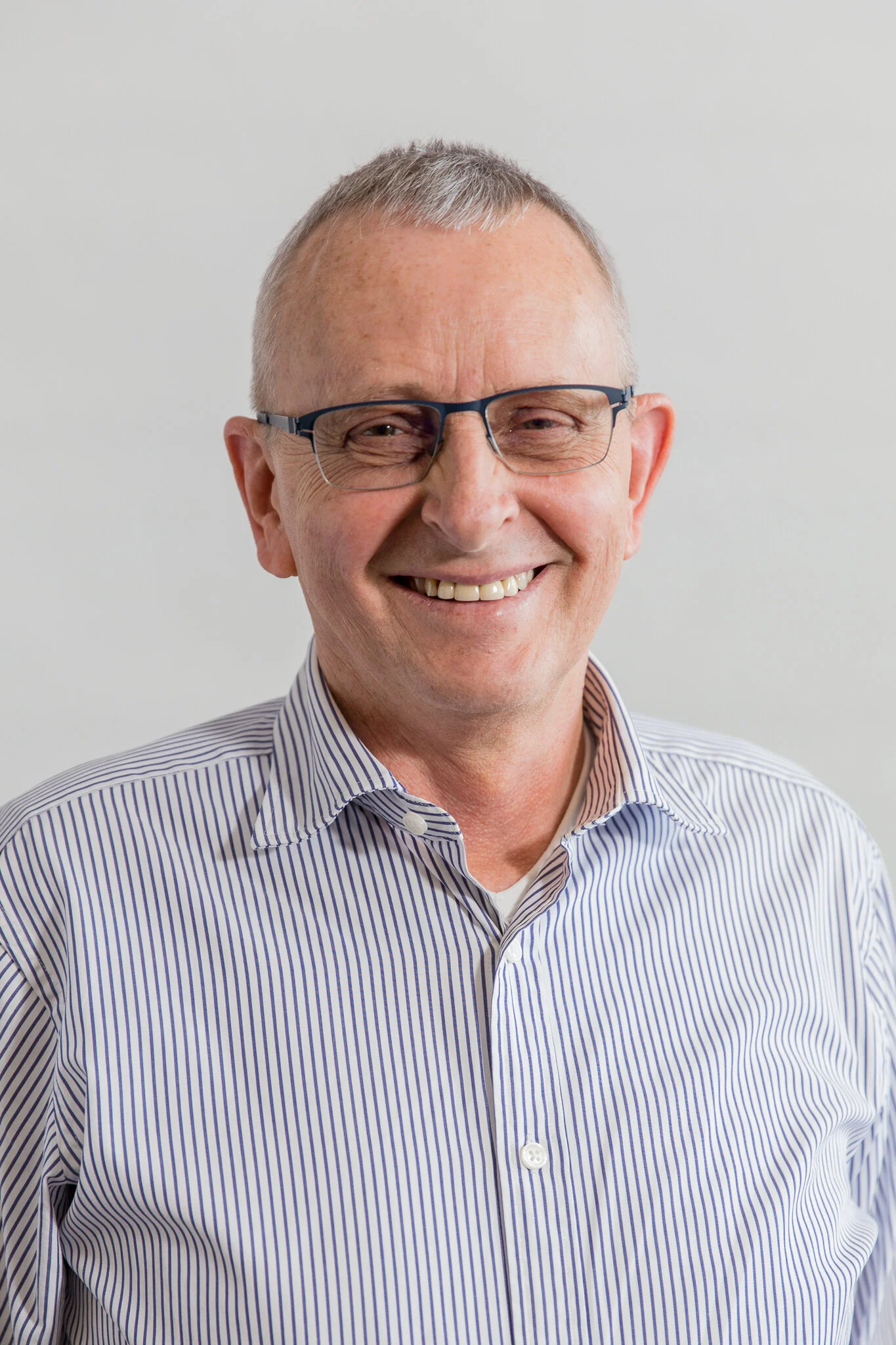 Headshot of a smiling middle-aged man with short gray hair and glasses, wearing a striped shirt, against a plain white background.