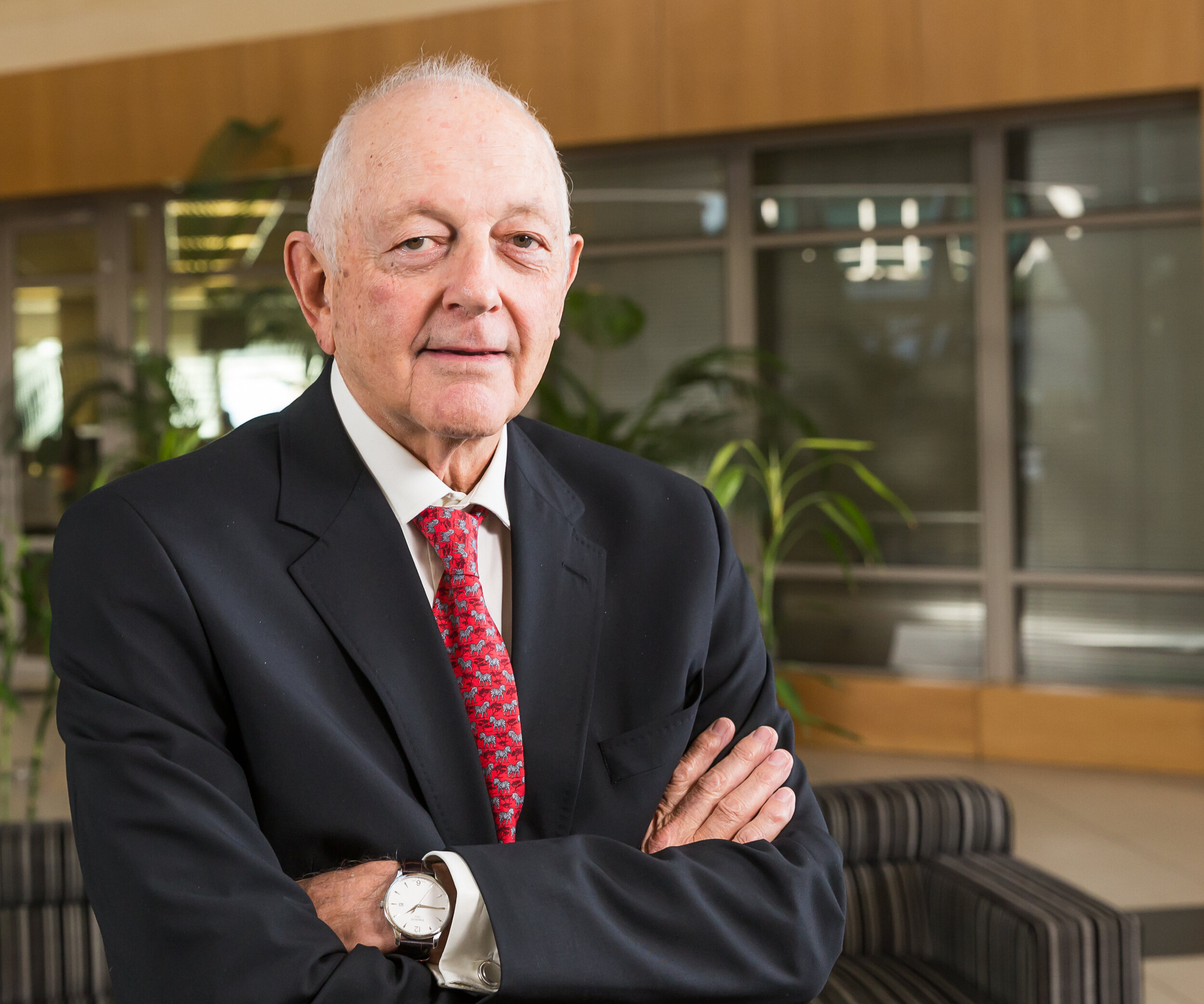 An elderly man in a black suit with a white shirt and a red patterned tie, standing indoors with his arms crossed, with plants and wooden furniture in the background.