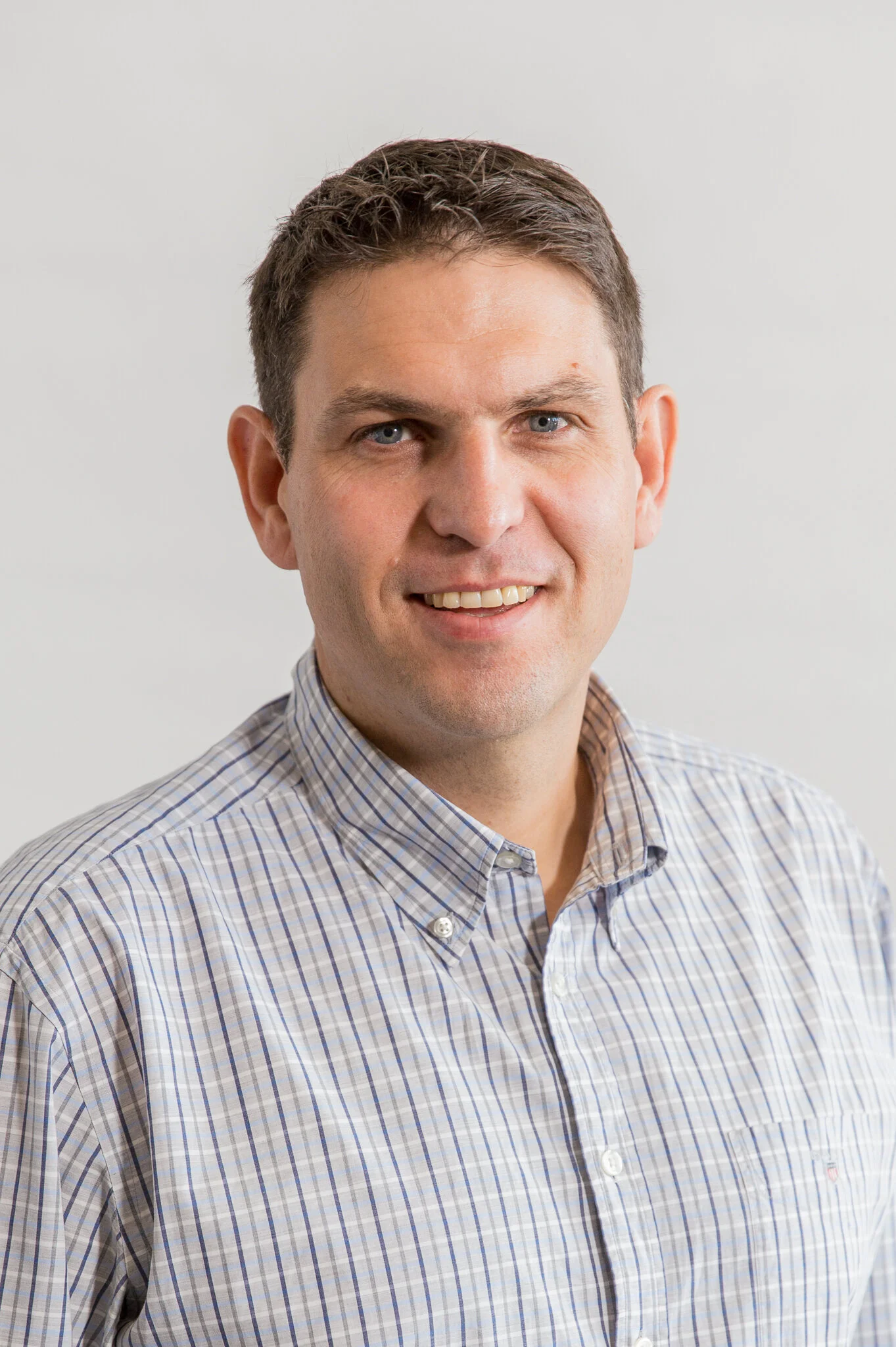 A middle-aged man with short dark hair, blue eyes, and a friendly smile wearing a light-colored, plaid button-up shirt in front of a plain light gray background.