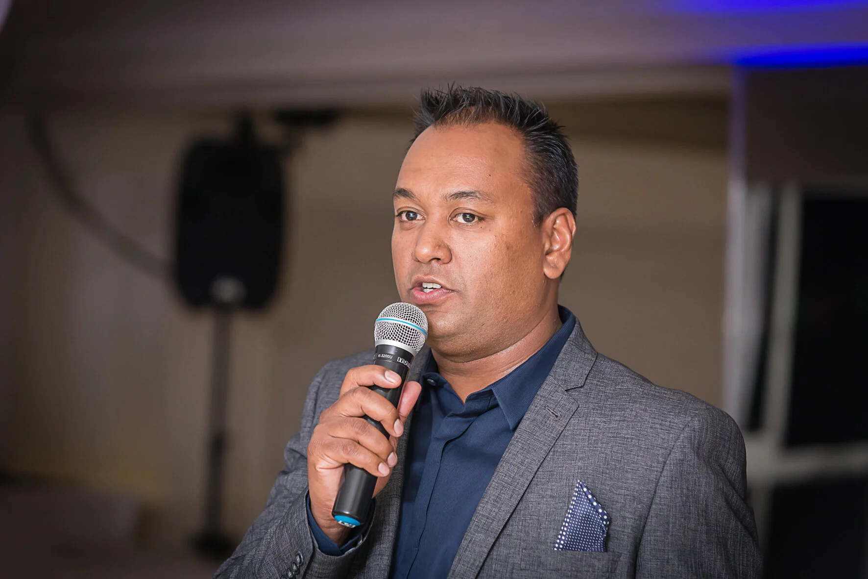 Man in gray suit speaking into a handheld microphone.