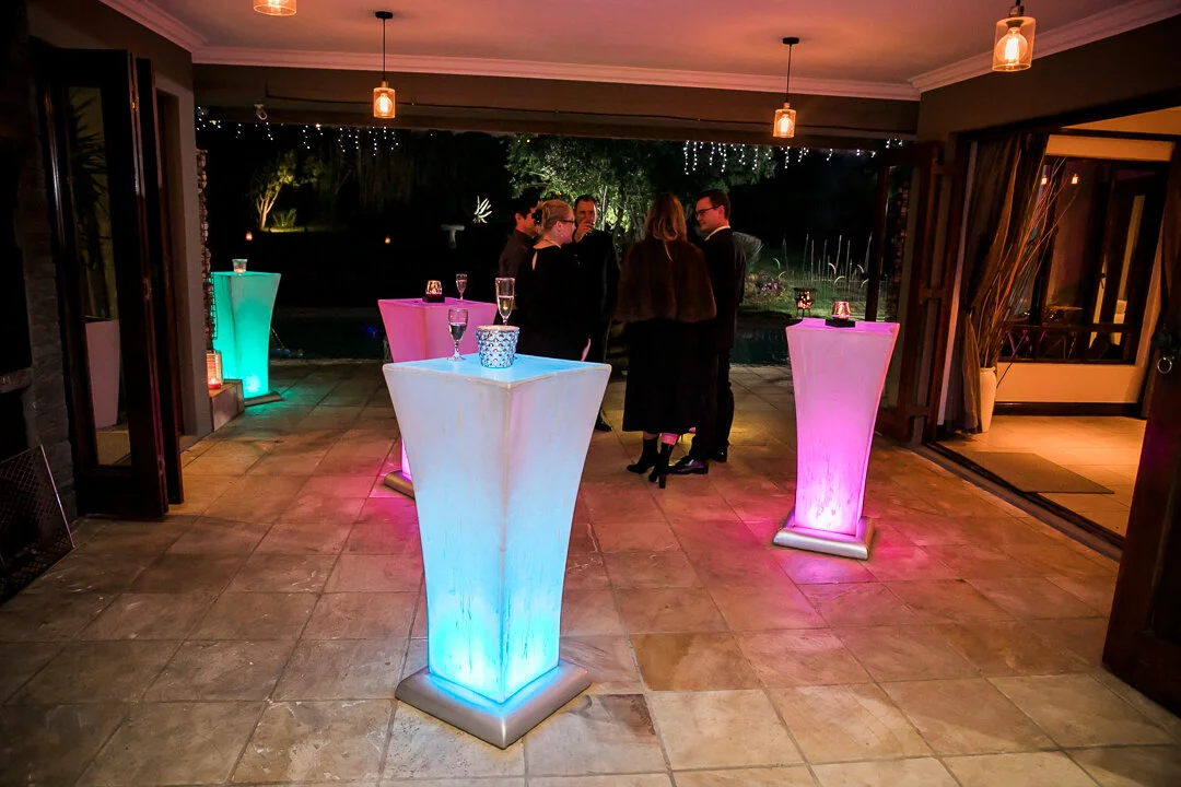 An indoor party scene with high-top tables illuminated with colorful lights in blue, pink, and green. Several guests are gathered outside on a patio, chatting, with string lights hung overhead and a garden visible in the background.