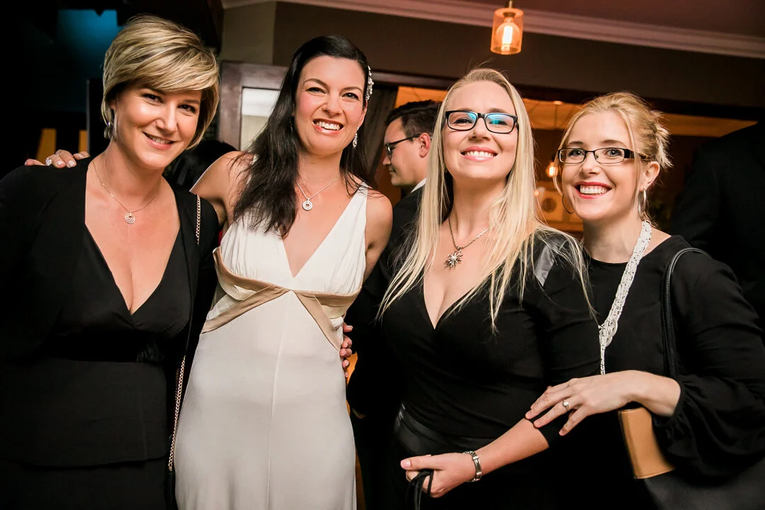 Four women smiling at an indoor party or event, dressed in elegant attire, standing close together, with a man in the background.
