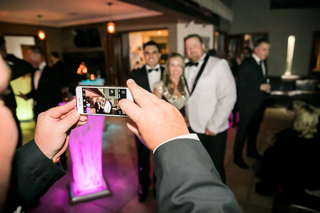 A person taking a photo of a group of people dressed in formal attire at a celebration or event in a dimly lit venue with pink lighting.