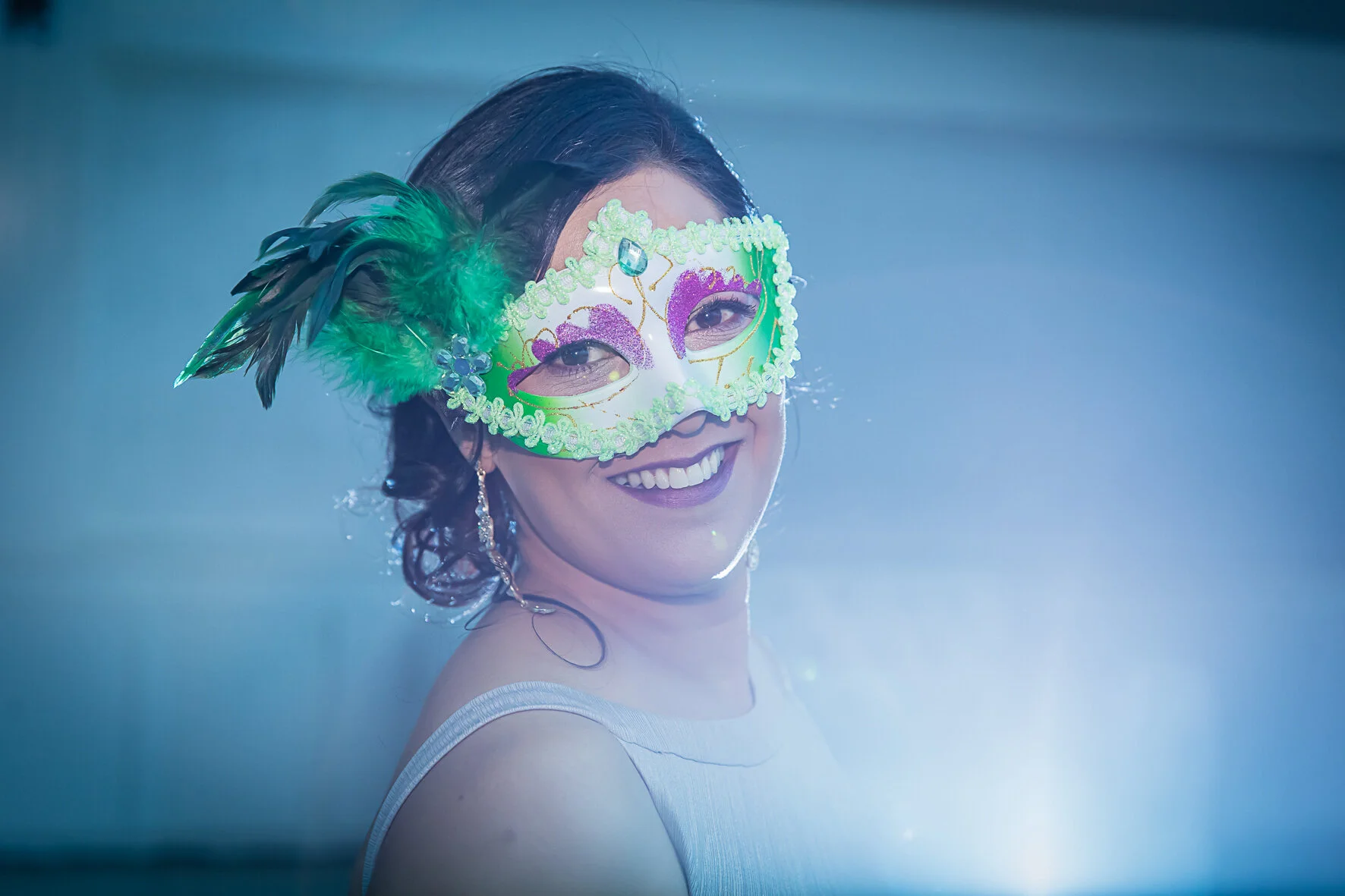 A woman wearing a colorful Mardi Gras mask with feathers, smiling, dressed in a white sleeveless dress, with earrings, illuminated by bright light and a blue background.