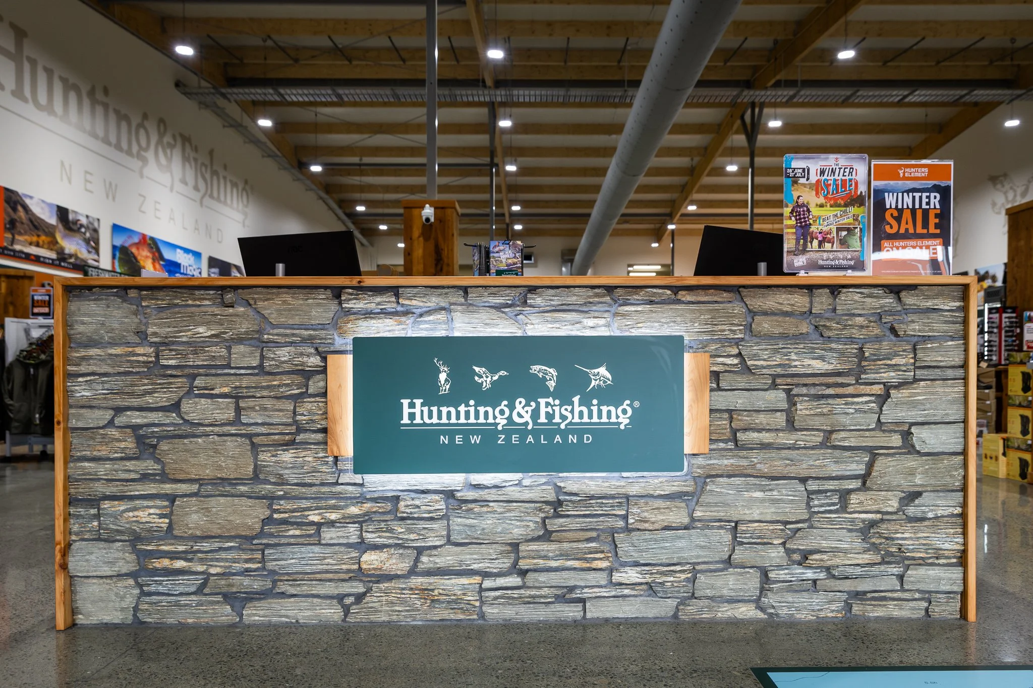 Hunting & Fishing New Zealand store counter with a stone facade and a green sign displaying hunting and fishing logo, surrounded by promotional posters for winter sale.