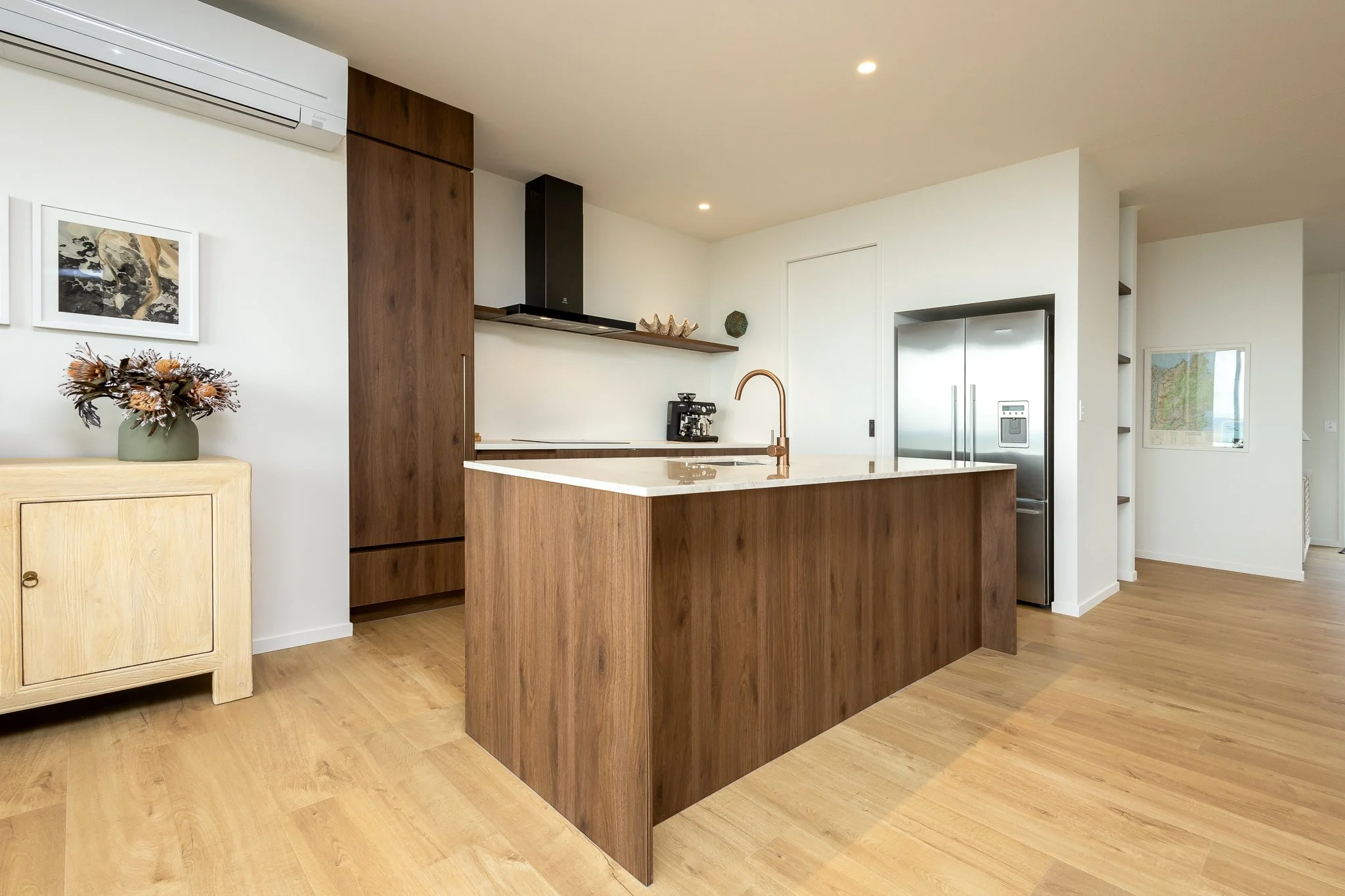 Modern kitchen interior photography with wooden cabinets and island, stainless steel refrigerator, black range hood, coffee maker, and decorative jars, in a bright space with wood flooring and minimal decor.