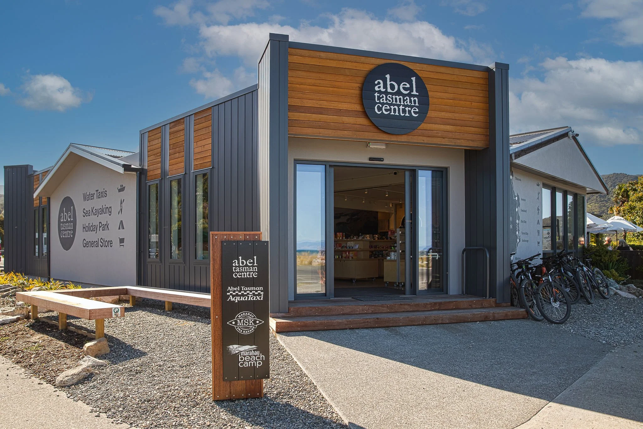 A building with a sign reading 'abel tasman centre' and another sign listing activities like water taxis, sea kayaking, holiday park, and general store. There are bicycles parked outside, and the sky is blue with some clouds.