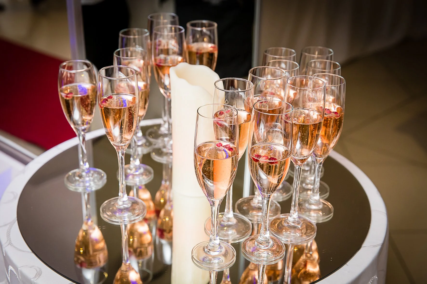 Multiple champagne flutes filled with a pink sparkling beverage, arranged on a round mirror table with a white candle in the center.