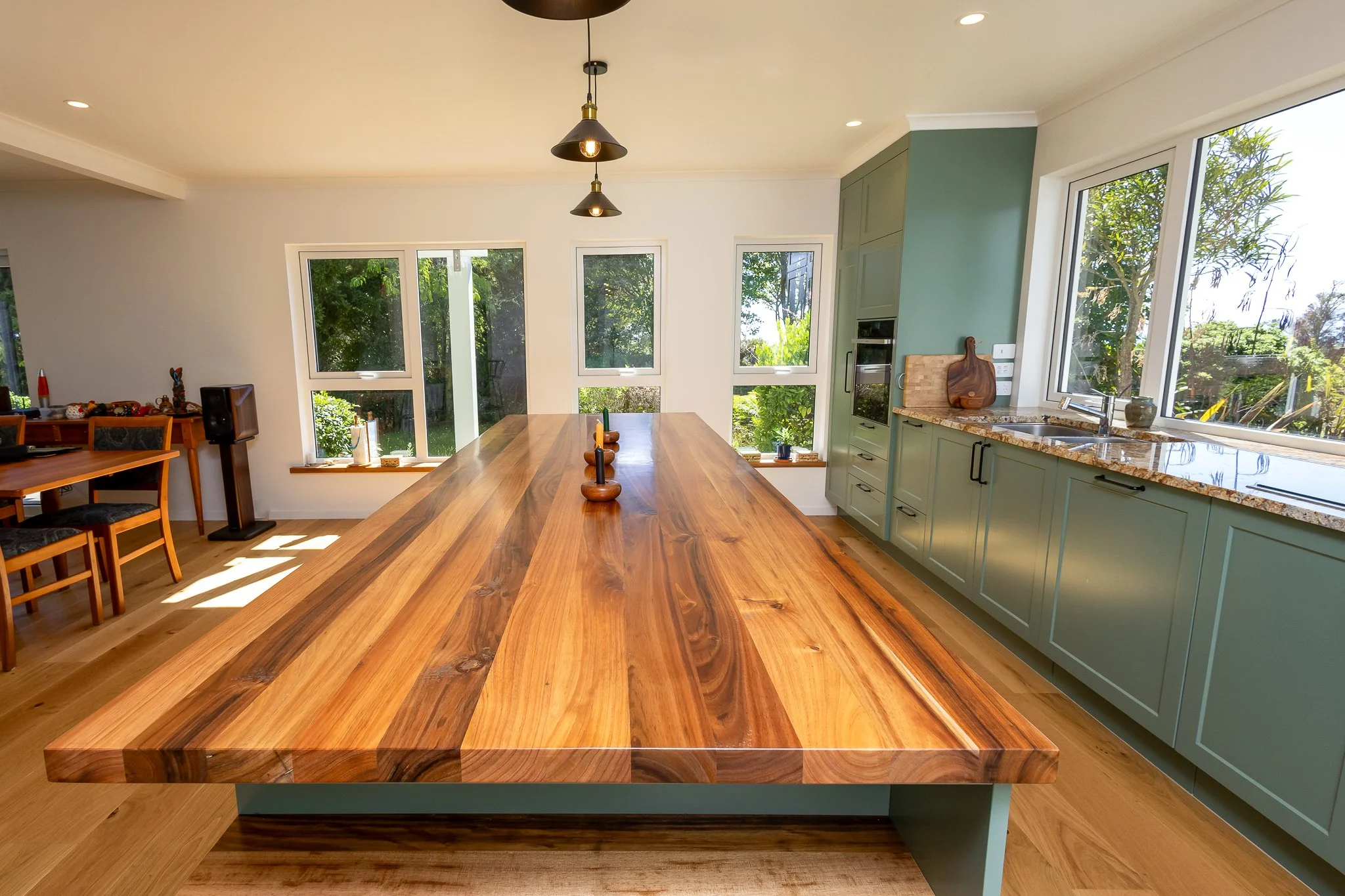A bright, modern kitchen with a large wooden dining table in the foreground, green cabinets, a marble countertop, and large windows overlooking a lush garden.