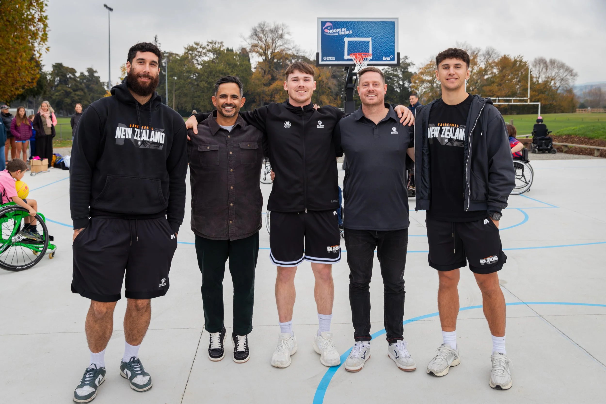 Group of five men standing on an outdoor basketball court, smiling, with basketball hoops and trees in the background. Two are wearing black athletic shorts and black sweatshirts that say 'New Zealand.' Others are in casual clothes.
