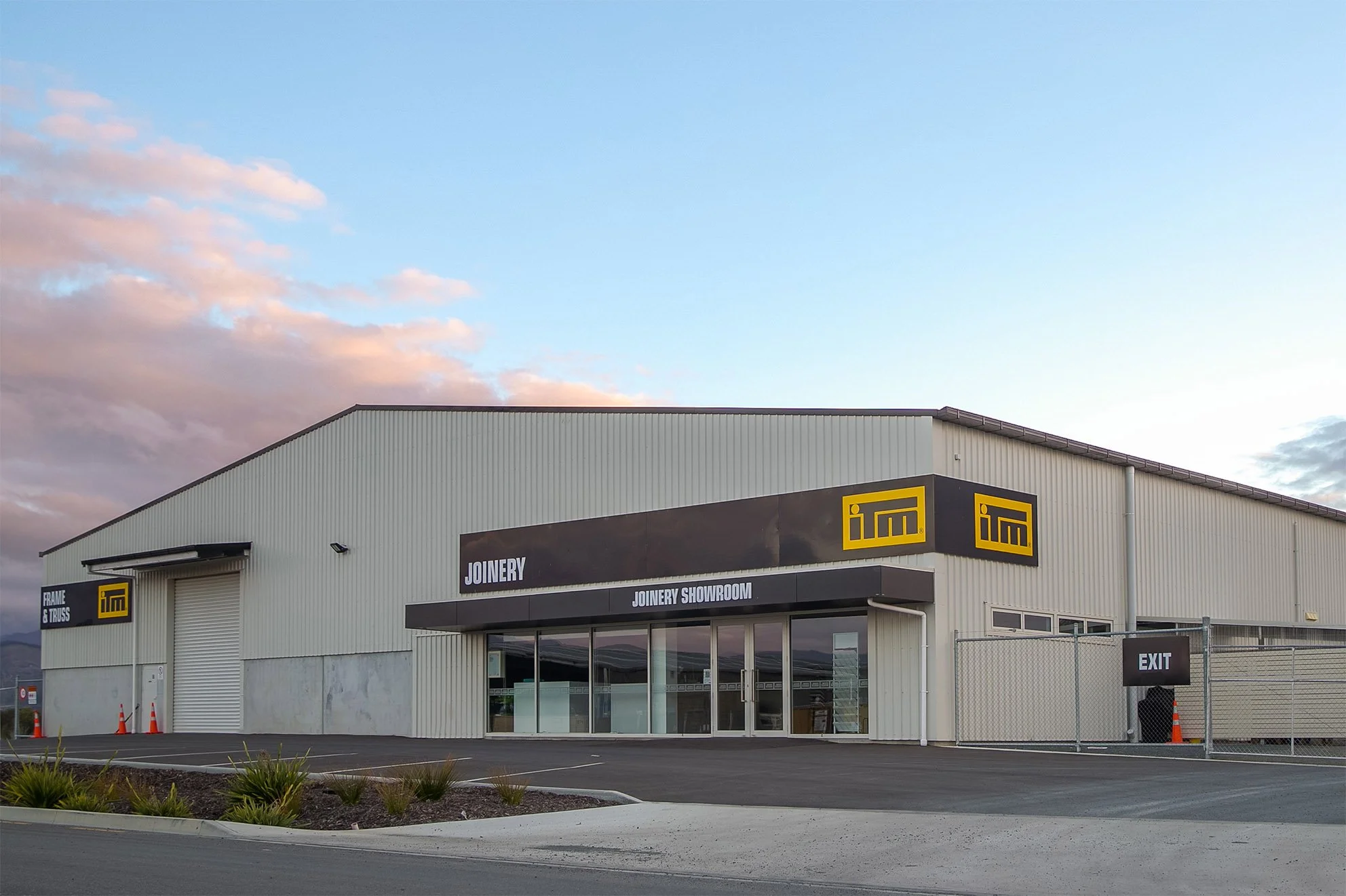 Exterior view of a large beige industrial building with signs indicating joinery and manufacturing services, parking lot in front, and a partly cloudy sky overhead.