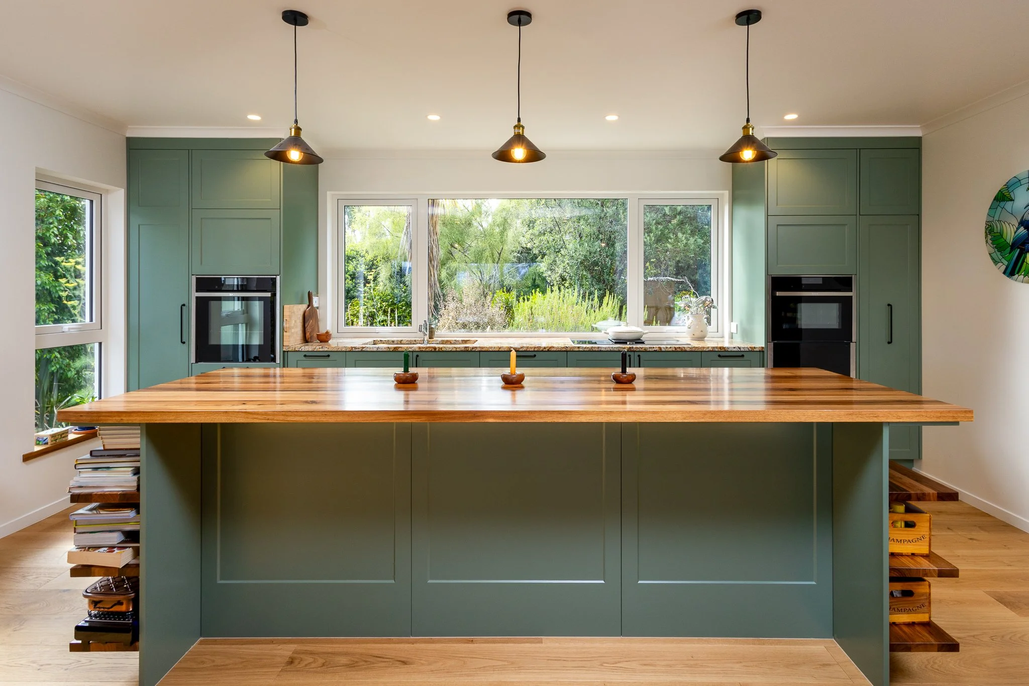 A modern kitchen with a large central island, green cabinetry, three pendant lights hanging from the ceiling, large windows showing greenery outside, and wooden countertops.