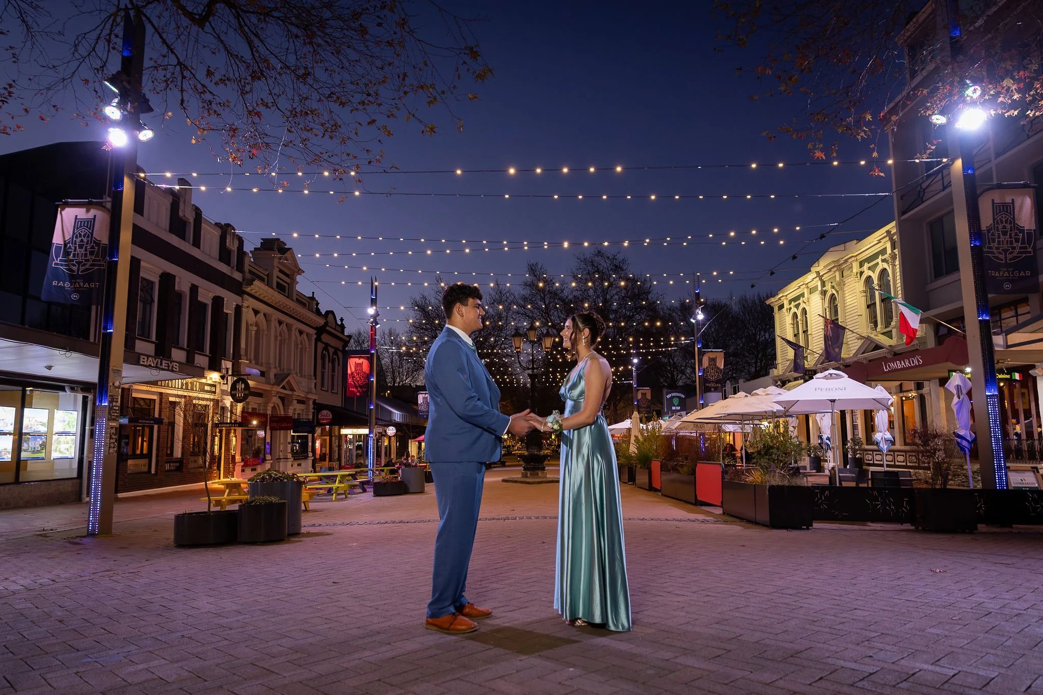 A couple dressed in formal attire stands under string lights on a city street at night, holding hands and looking at each other, with shops and outdoor umbrellas behind them.