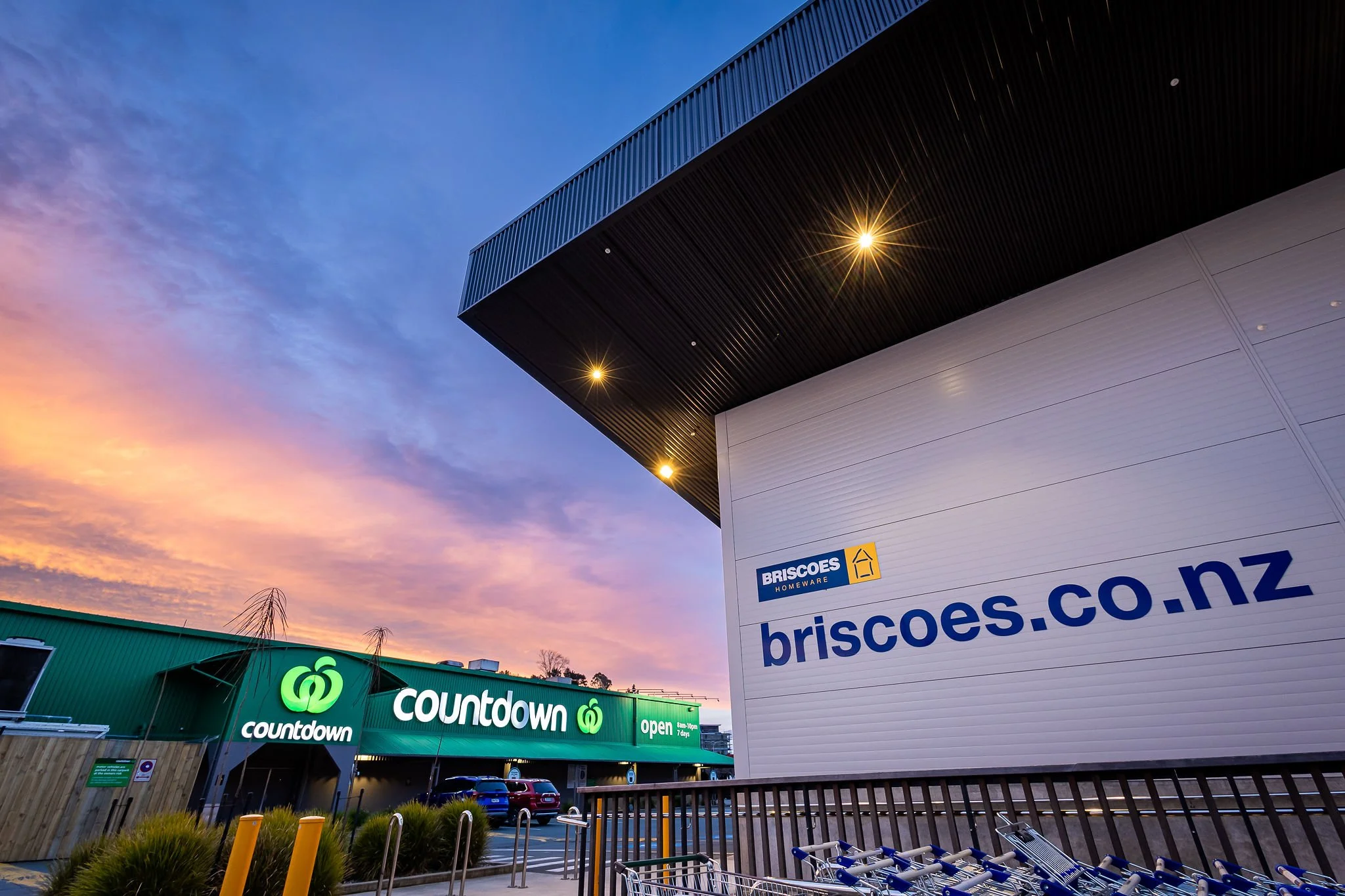 Storefronts at sunset with Briscoes Homeware sign on a white building and Countdown grocery store with green signage beneath a colorful sky.