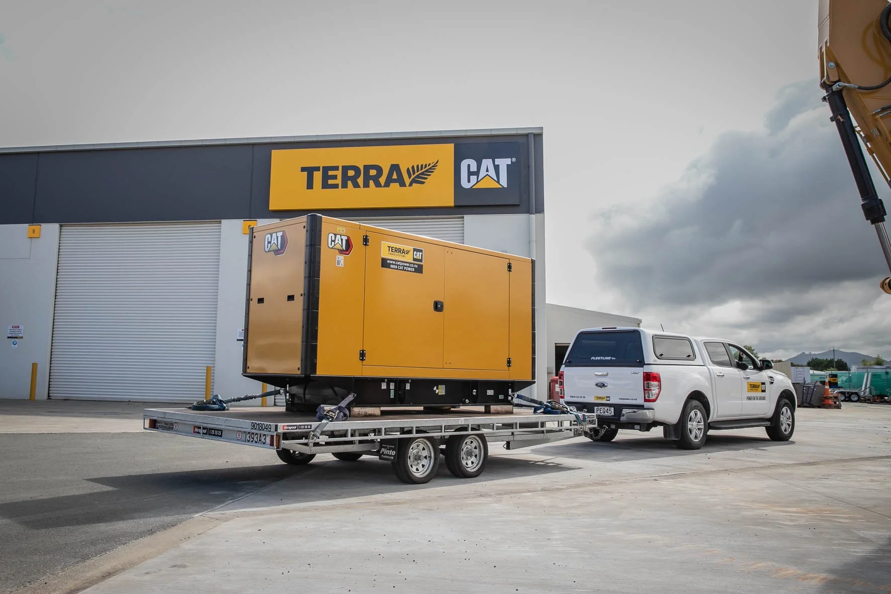 A construction equipment truck parked outside a building with a large yellow CAT generator on a flatbed trailer, with a white pickup truck attached, near a warehouse with a Terra CAT sign and cloudy sky.