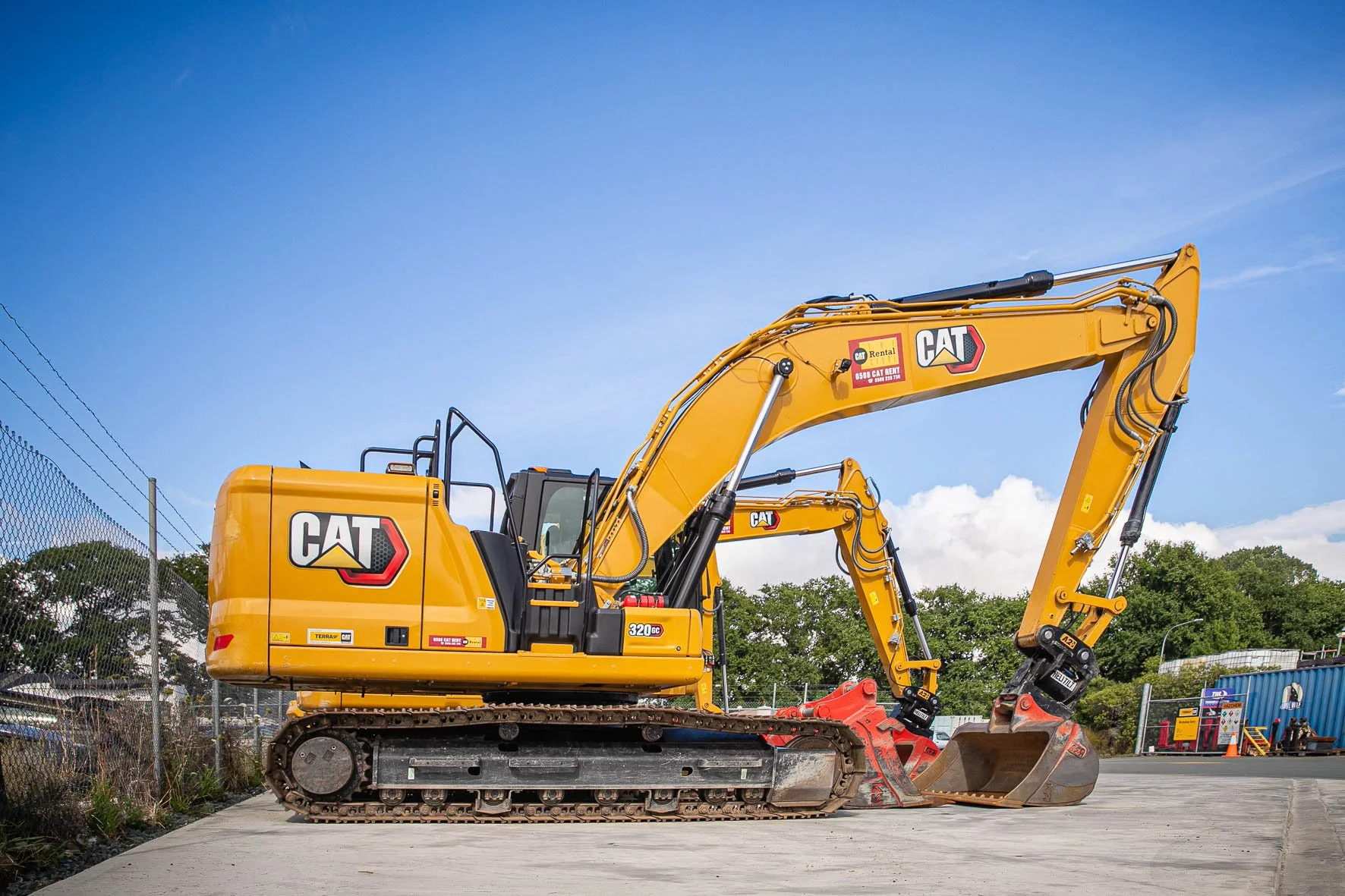 A yellow Caterpillar excavator with black tracks parked outdoors on a construction site, with a chain-link fence, trees, and a blue sky with some clouds in the background.