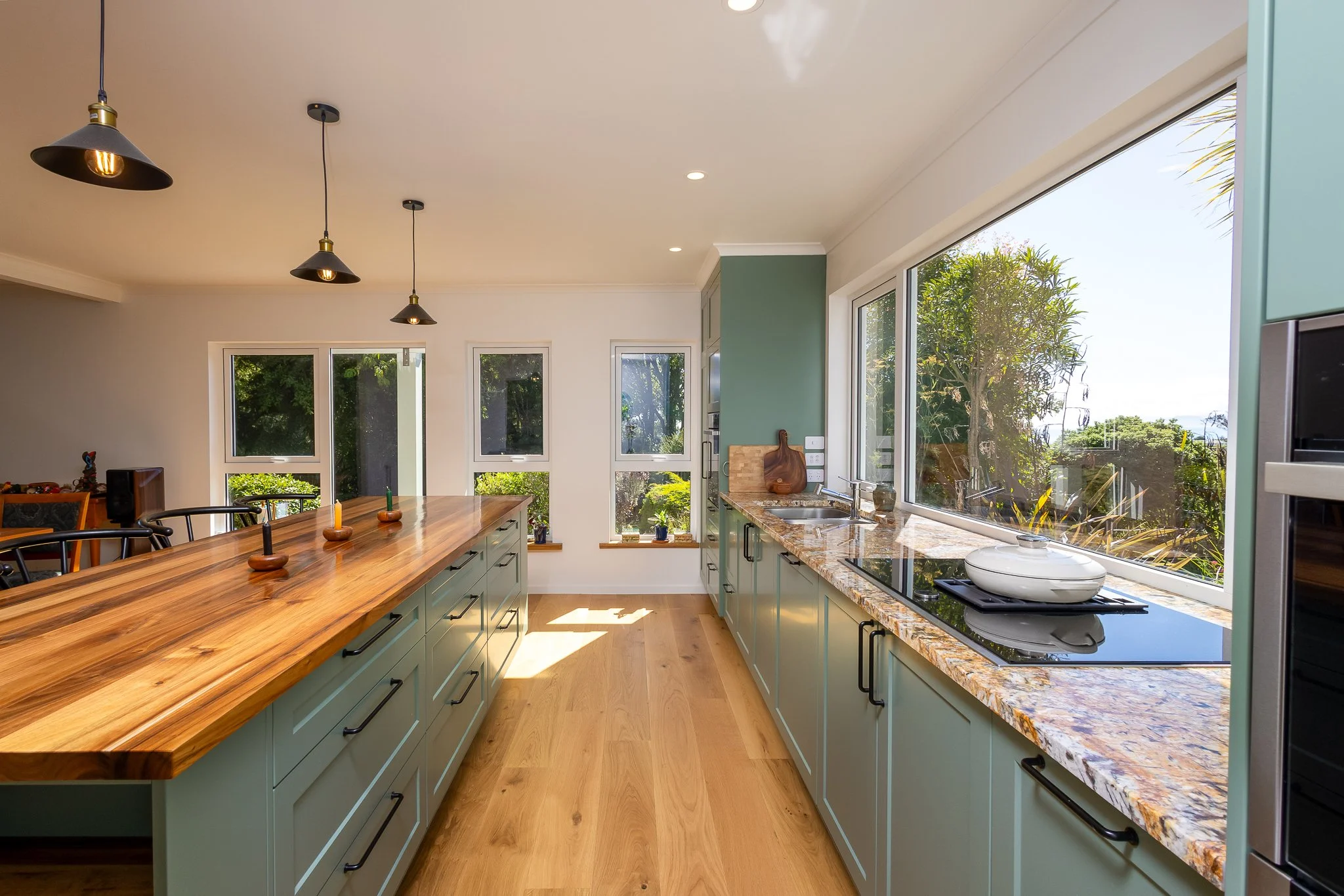A modern kitchen with green cabinets, a large wooden island, and a granite countertop with a built-in stove. Large windows reveal a view of trees outside, and pendant and recessed lighting illuminate the space.