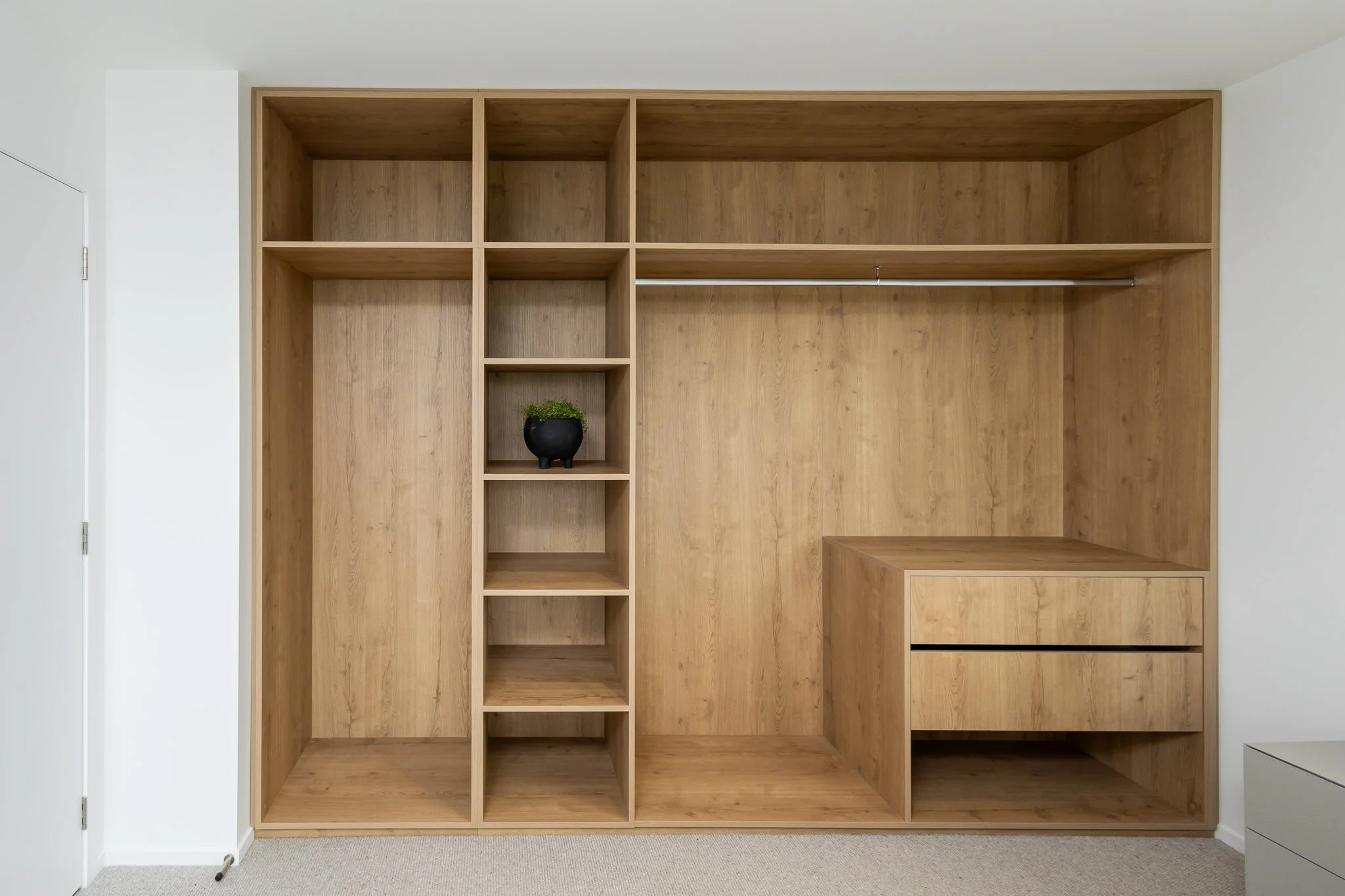 Empty wooden closet with open shelves and a hanging rod, a small black pot with a green plant inside, and a closed drawer at the bottom right.