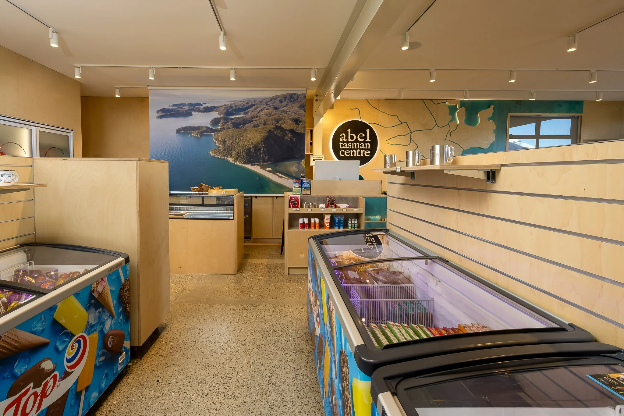 Interior view of a gift shop with ice cream freezers and shelves, a large mural of a lake and mountains on the back wall, and a sign that says 'abel tasman centre'.