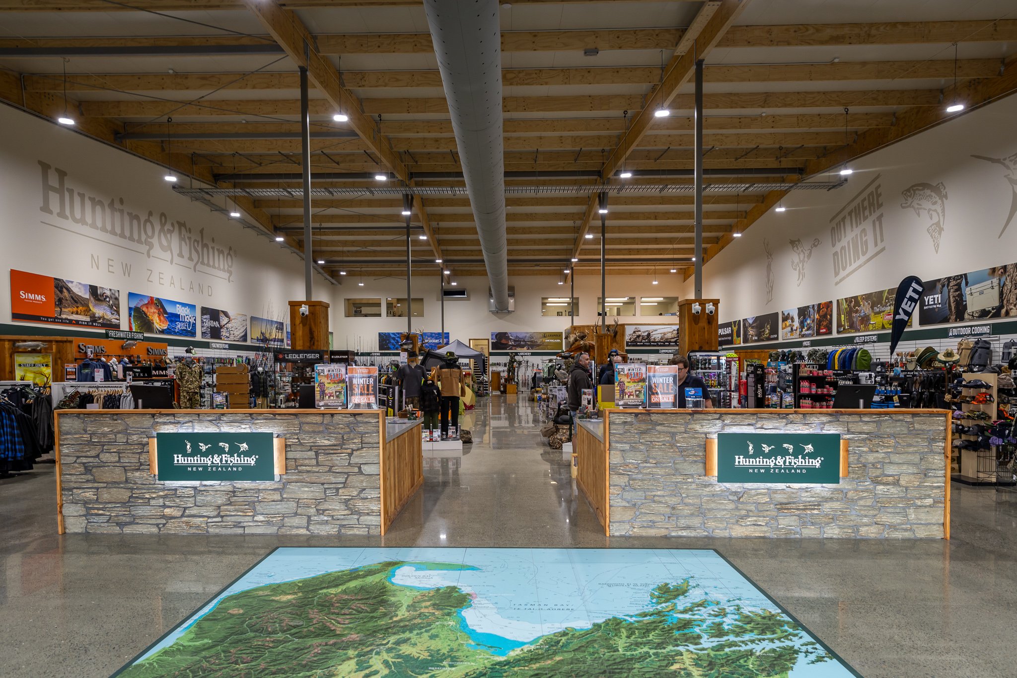 Interior of a hunting and fishing store with displays of outdoor gear, a large map on the floor, and signage that reads 'Hunting & Fishing New Zealand' and 'Out Here Doing It'.
