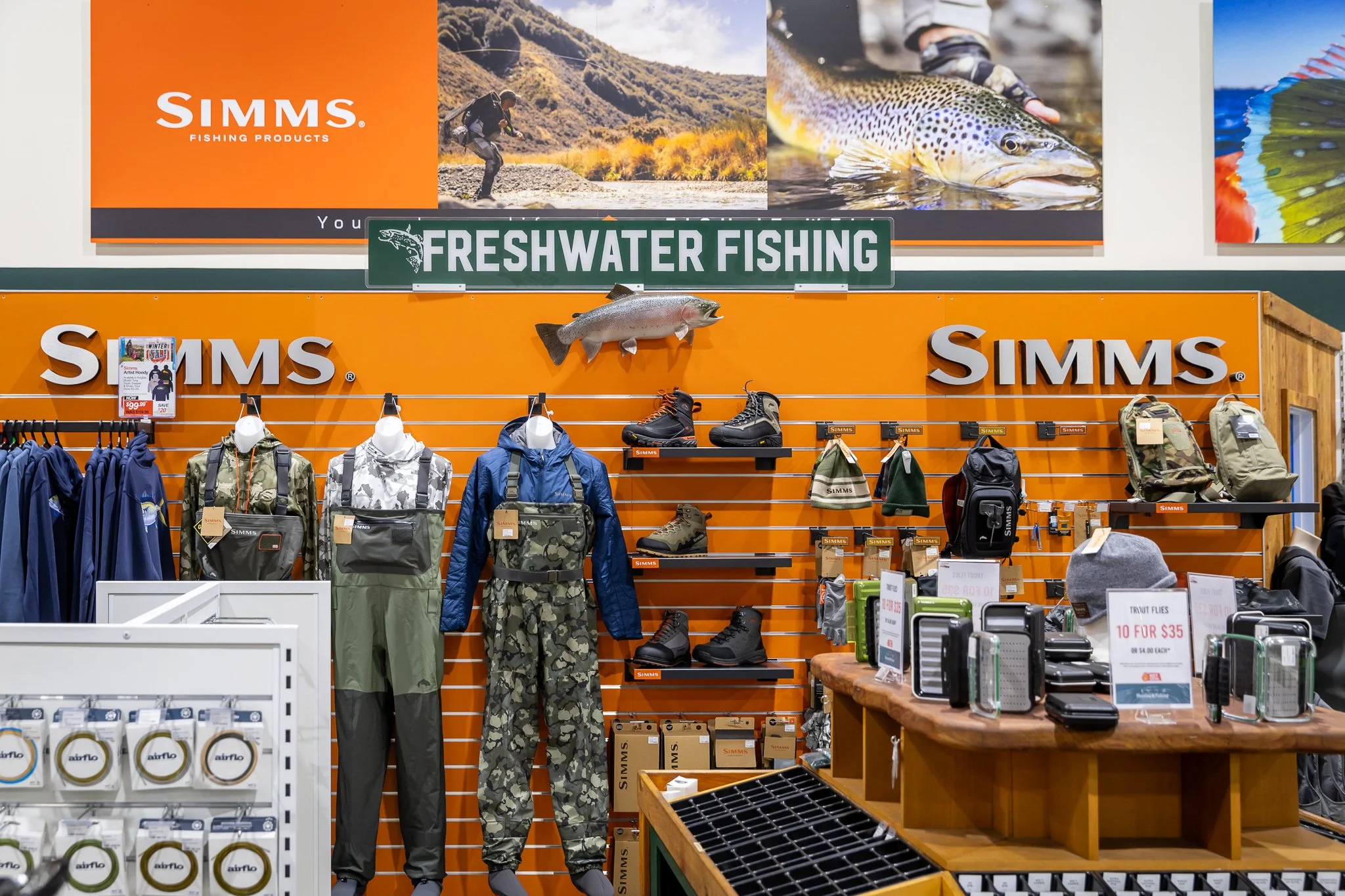 Store display with outdoor and fishing gear, including jackets, boots, and backpacks, featuring the Simms brand, with a sign for freshwater fishing and a mounted fish.