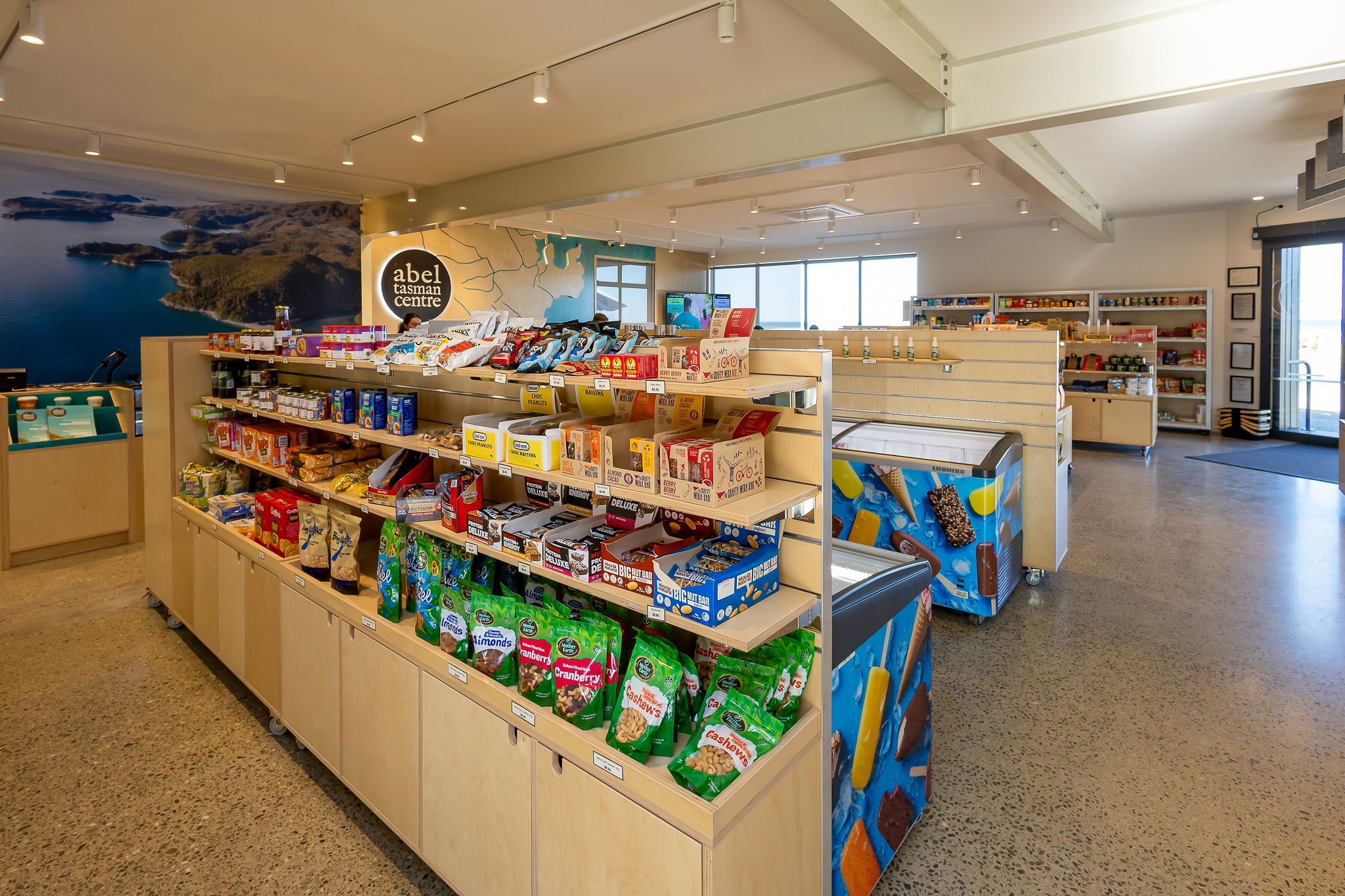Interior view of a store, with shelves stocked with snacks such as chocolates, nuts, and bars, and a mural of Tasmanian landscapes on the wall behind the shelves.