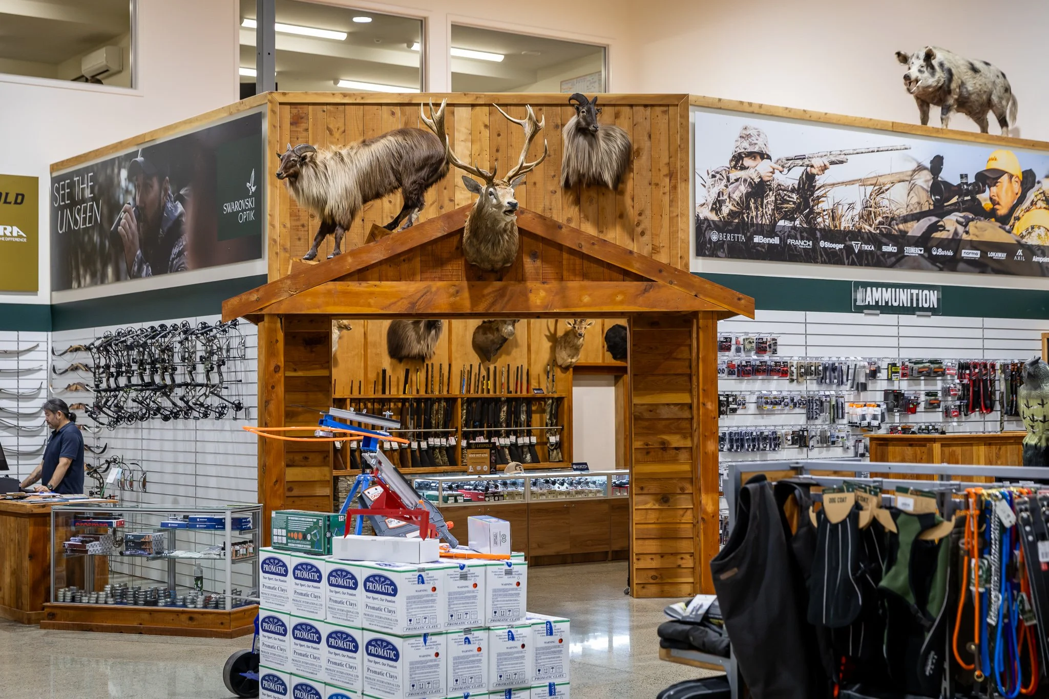 Display inside a store with mounted animal heads of a moose, a ram, and a deer on a wooden wall. Shelves below hold hunting rifles and ammunition. Various brands and hunting gear are visible.