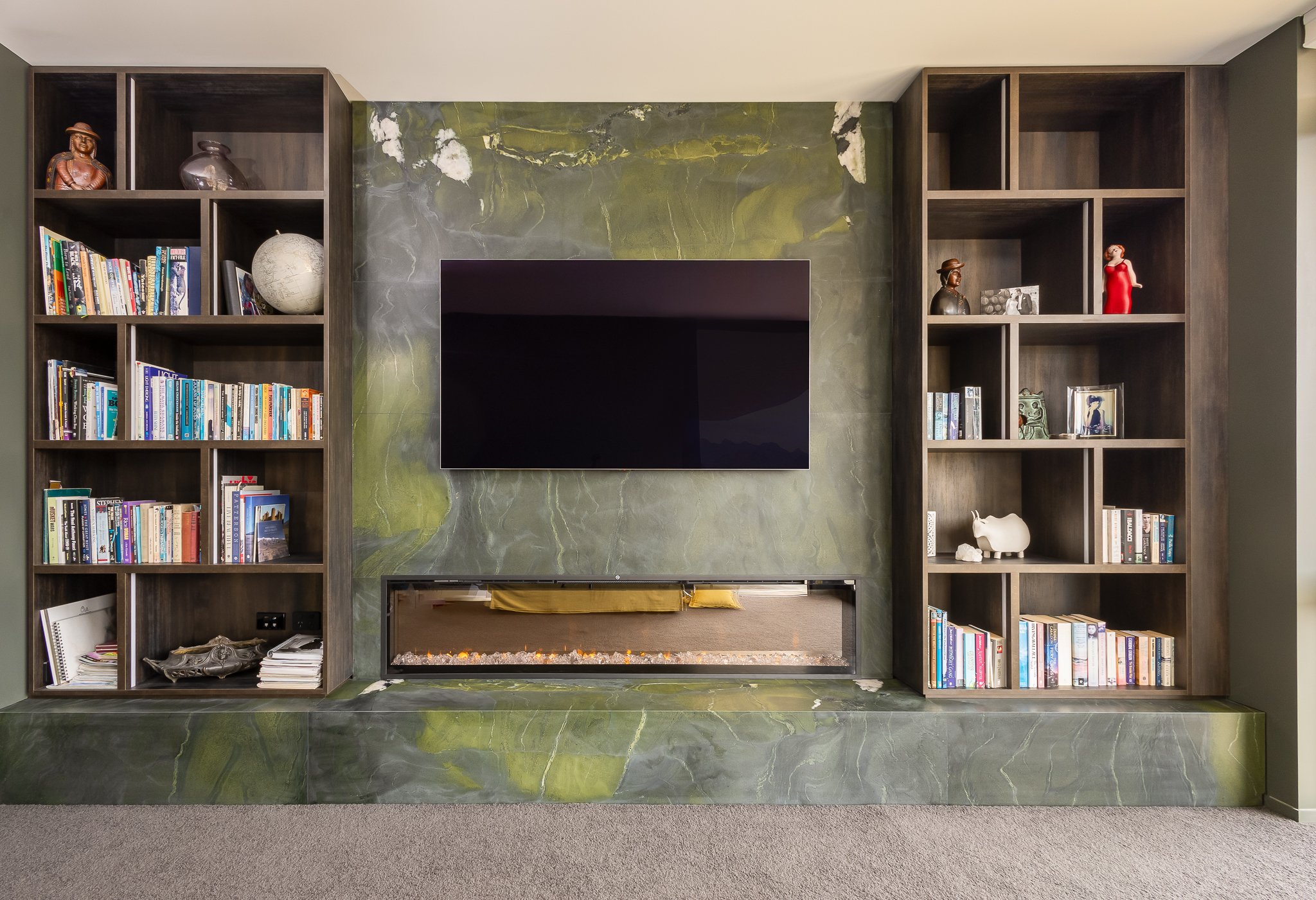 Living room wall with dark wood bookshelf on either side, a green marble fireplace with a black TV mounted above, and decorative items including books, sculptures, and framed photos.