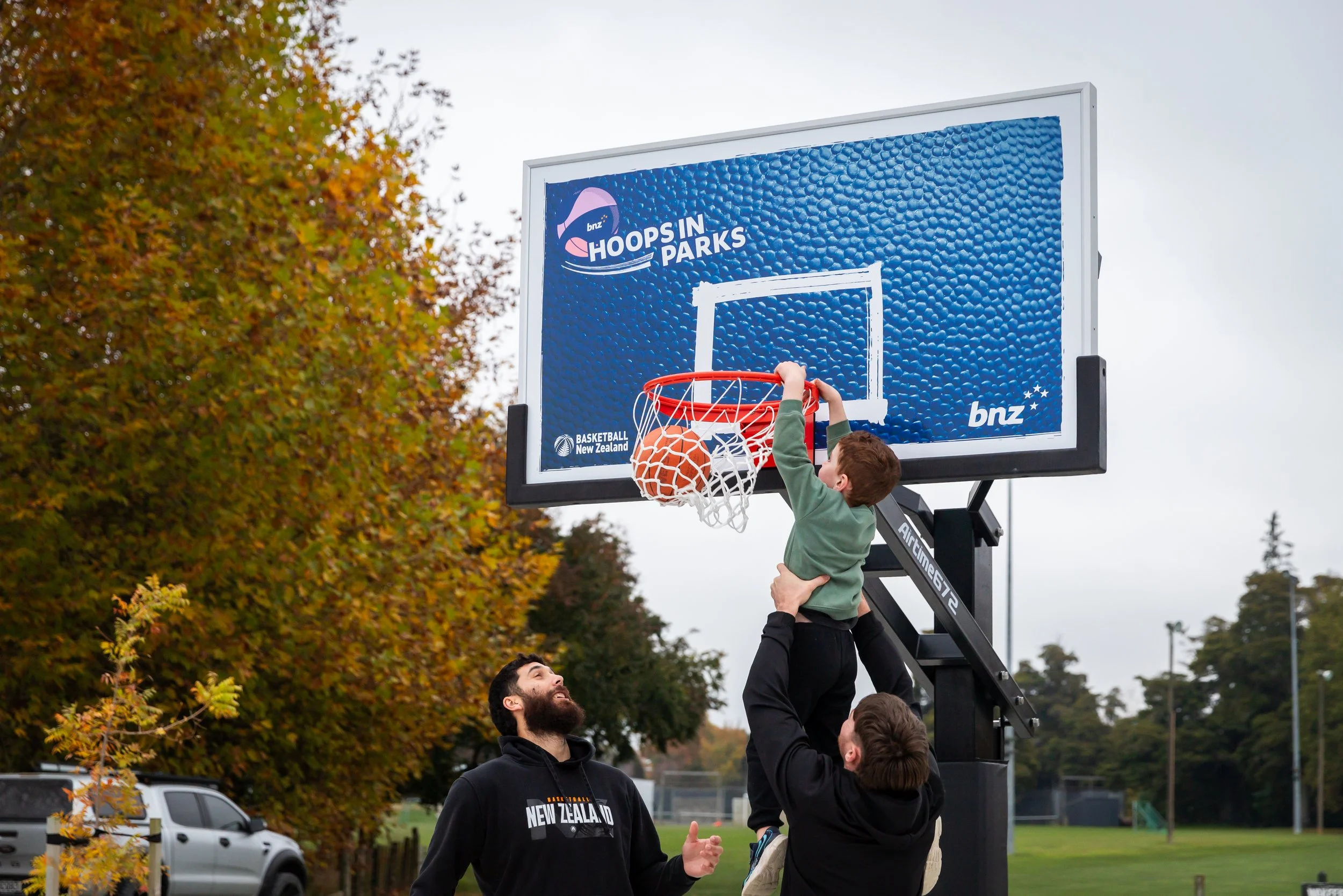 Children playing basketball outdoors during autumn with a backboard displaying the 'Hoops in Parks' and 'Basketball New Zealand' logos. One child is shooting a basketball into the hoop, while others are watching and smiling.