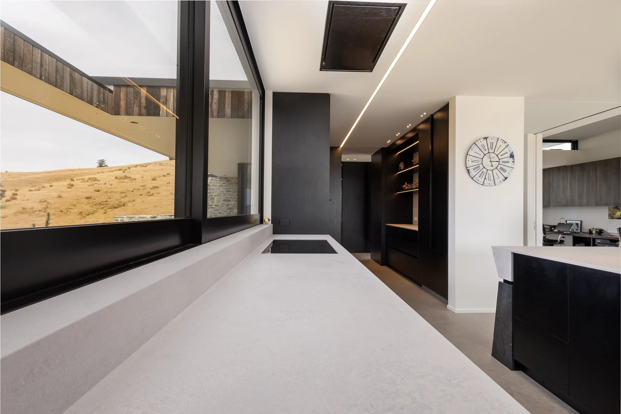 Modern kitchen interior photography with large window overlooking dry landscape, white countertop, black accents, wall clock, and built-in shelves.