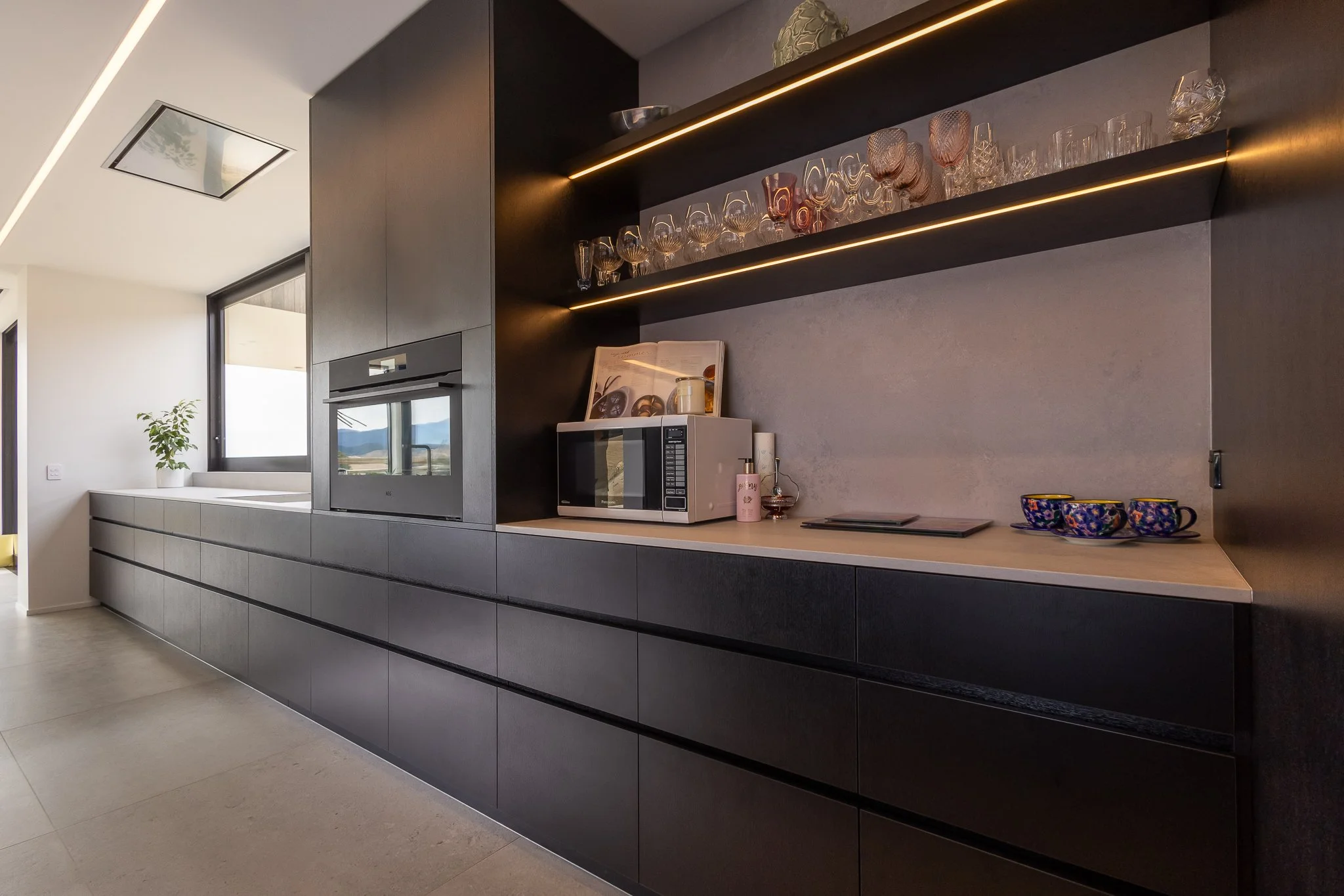 Modern kitchen interior photography with black cabinets, open shelves with glassware, microwave, and decorative bowls, and a window with a view of mountains.
