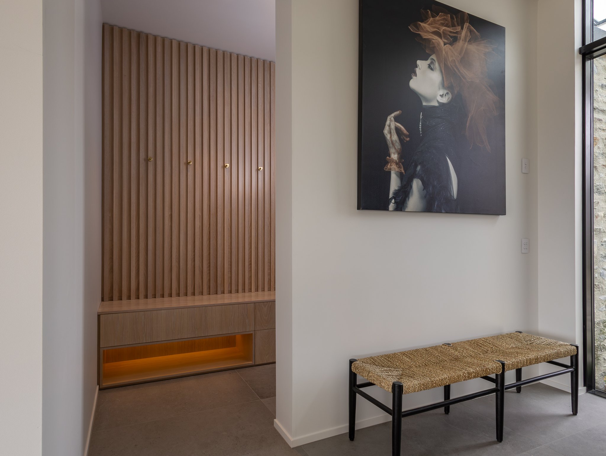 Modern interior entryway with wooden panel closet, large black and white artistic poster of a woman with voluminous hair, a woven bench, and a glass door with a view of the outdoors.