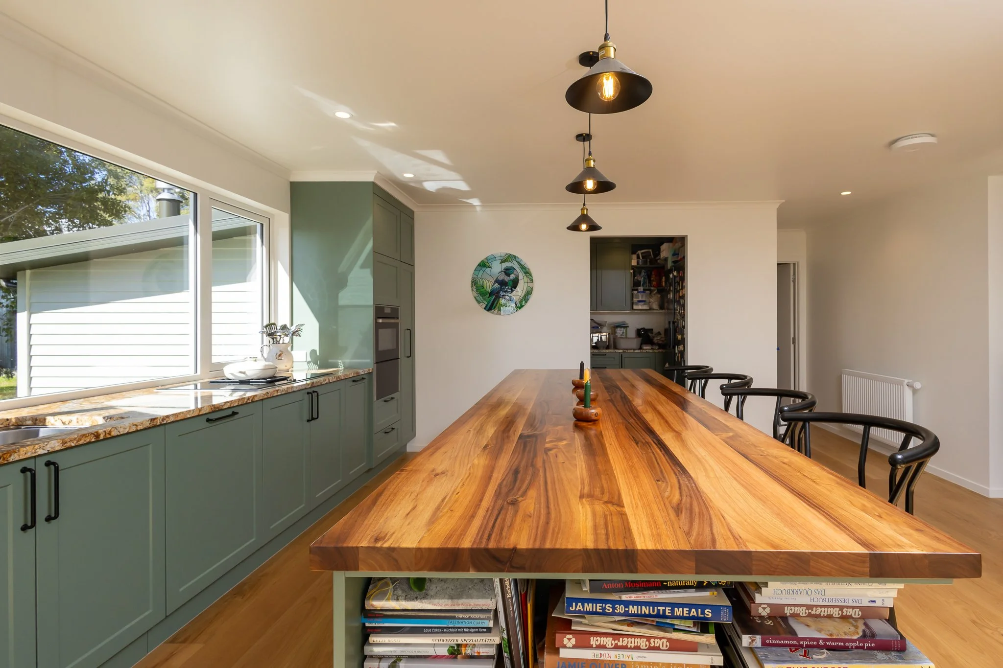 Open-concept kitchen with green cabinets and a large wooden dining table, pendant lights hanging from the ceiling, large window with a view outside, and a shelf with cookbooks underneath the table.