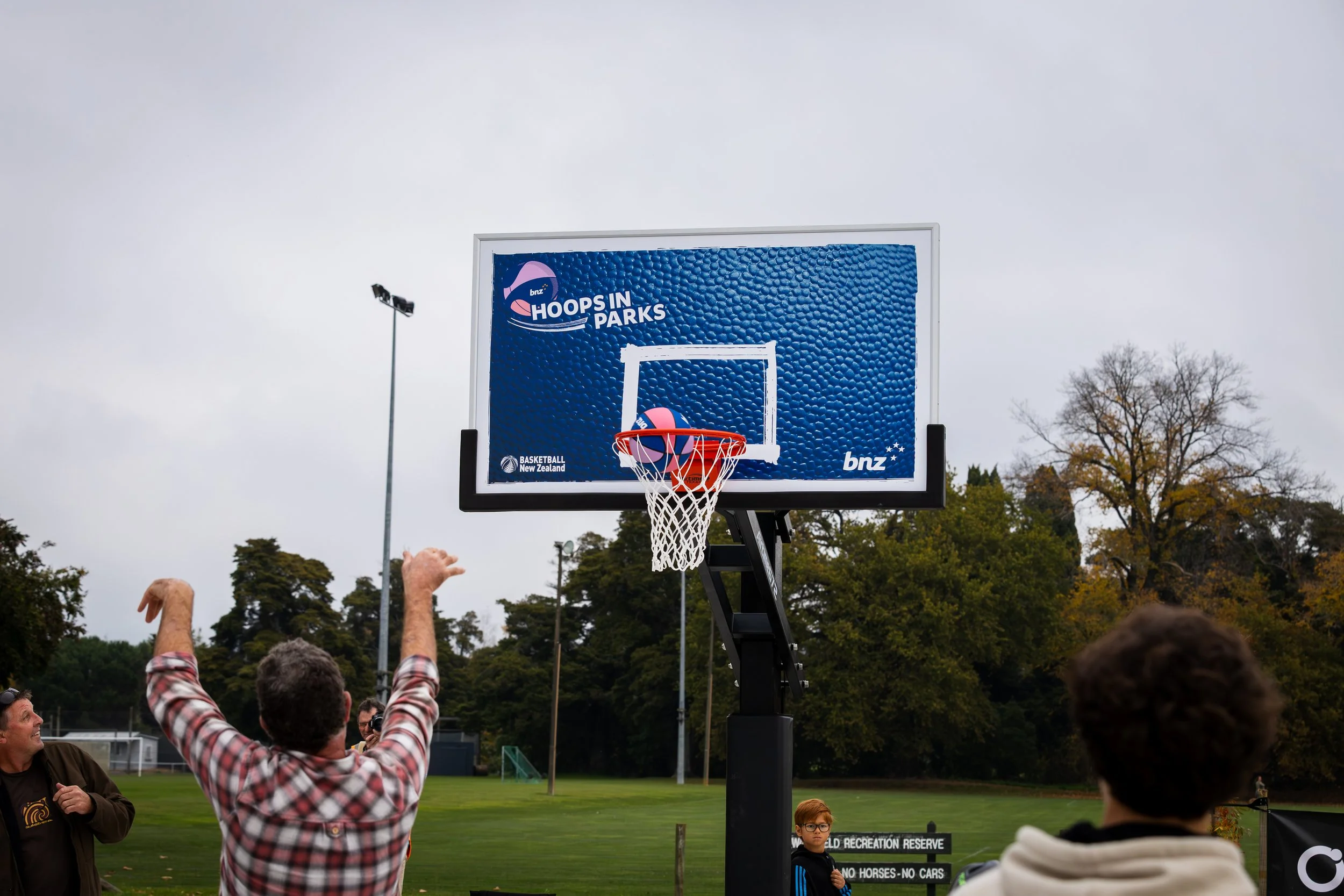 People playing basketball outdoors on a court with a large digital scoreboard displaying a logo and basketball graphics, some spectators watching the game, trees in the background, and overcast sky.