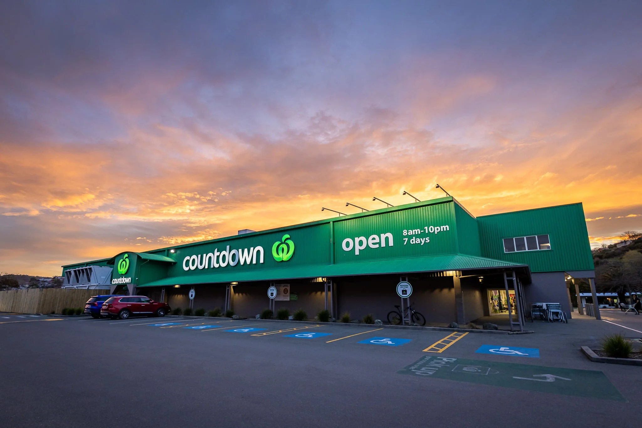 Exterior of a green Countdown store at sunset, with parking lot in the foreground and blue sky with clouds in the background.