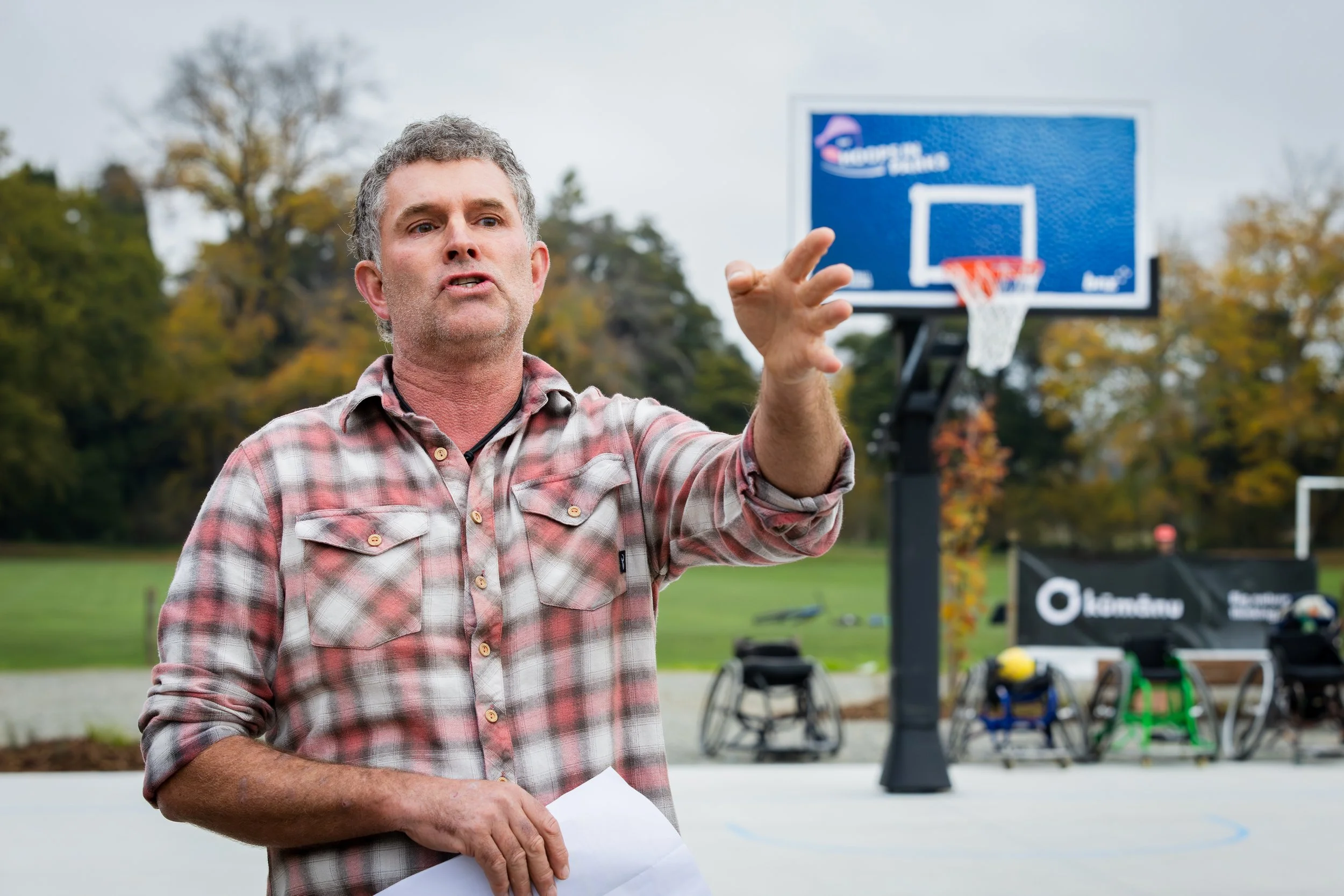 A man speaking outdoors near a basketball hoop, holding papers in his hand, with trees and sports equipment in the background.