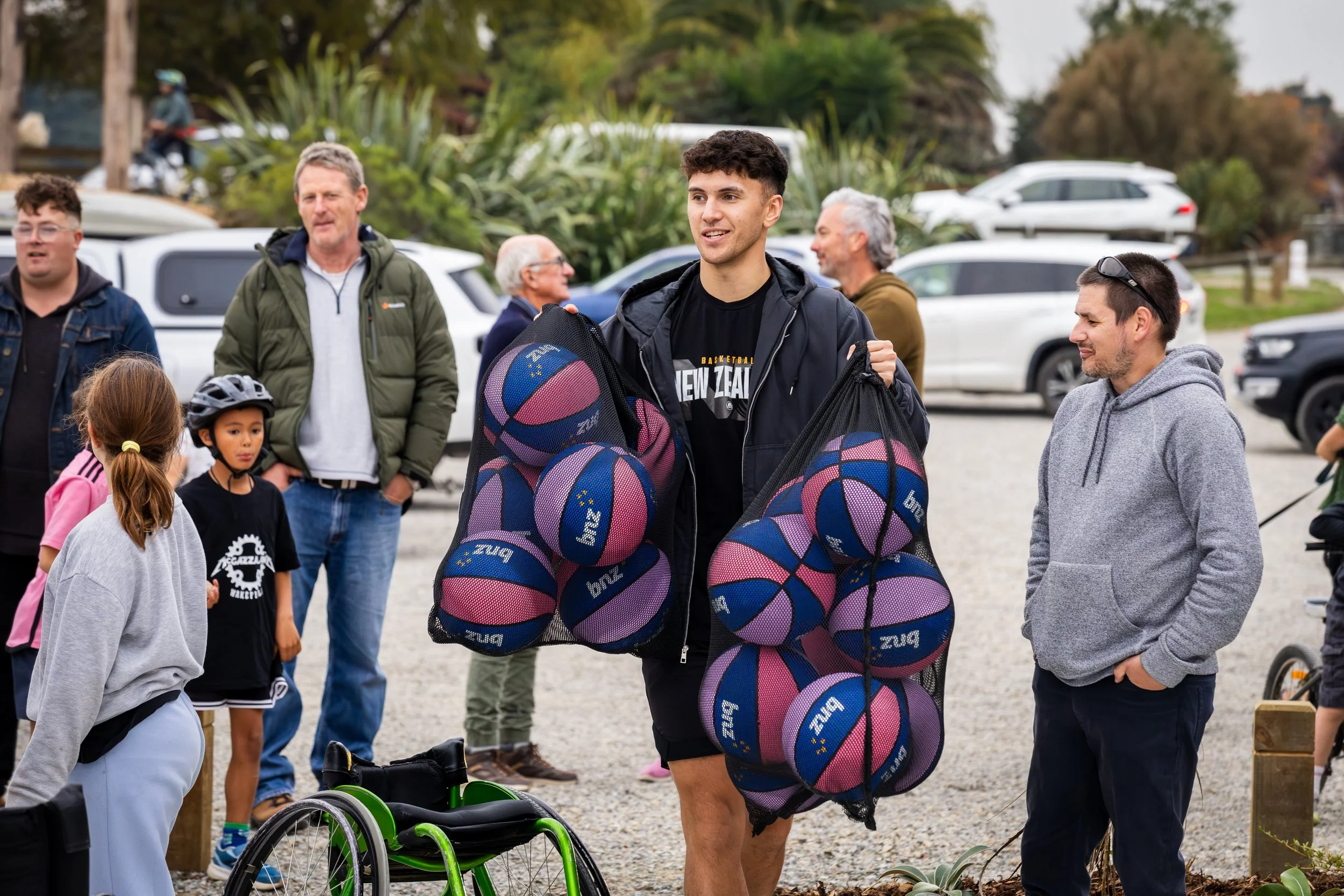 Group of people, including young man carrying multiple basketballs, gathered outdoors in a parking lot surrounded by trees and parked cars.