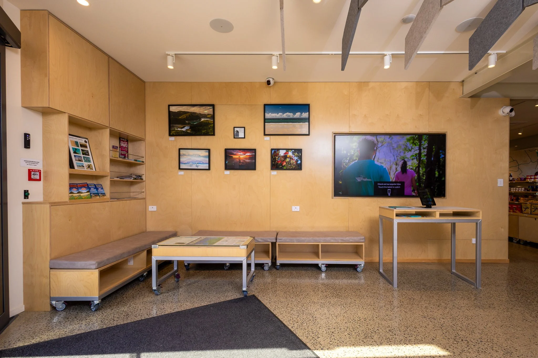 An indoor art gallery with framed landscape photographs on a wooden wall, featuring a seating area with benches on wheels, and a large digital display screen showing an outdoor scene with children.