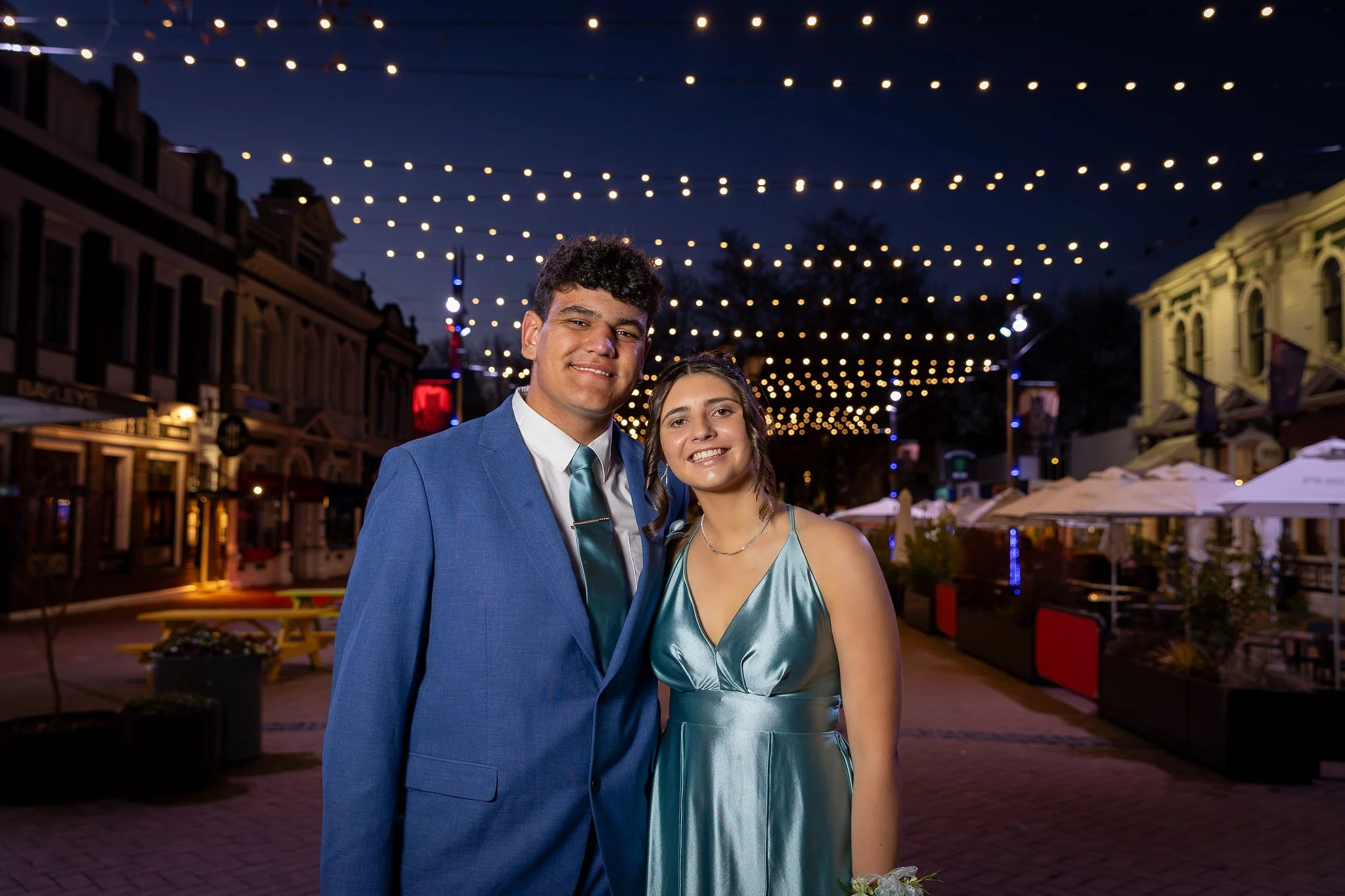A young man in a blue suit and a young woman in a matching blue dress are smiling and standing close together in an outdoor area decorated with string lights at night. There are buildings and outdoor furniture in the background.