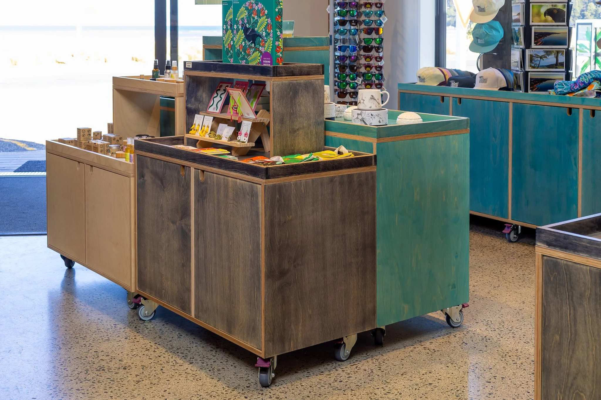 A retail display stand on wheels containing various souvenirs and accessories, including sunglasses, hats, postcards, and small decorative items, inside a brightly lit store with large windows.