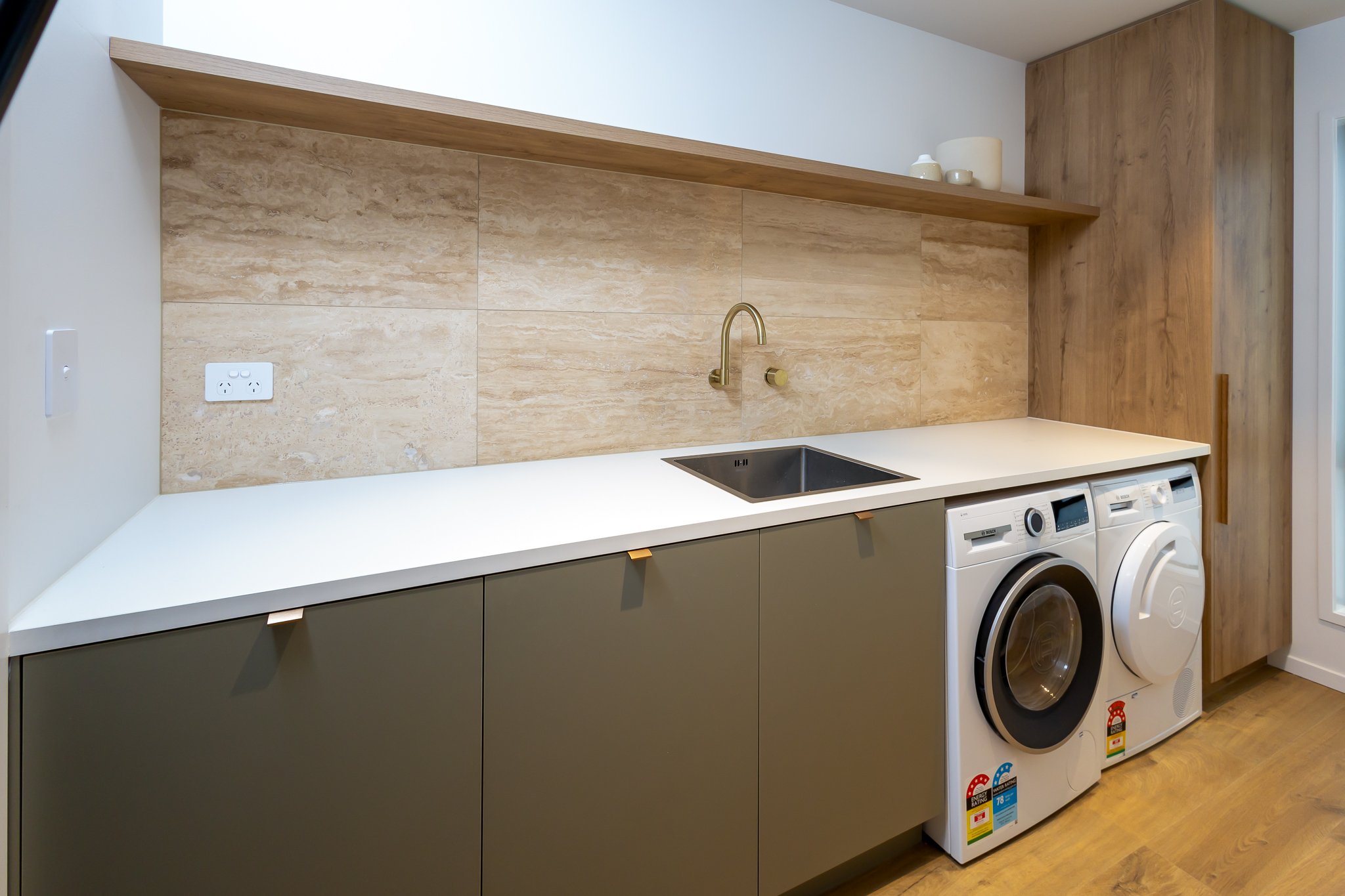 Modern laundry room with beige tiled backsplash, a gold faucet over a small black sink, gray lower cabinets, and a washing machine with laundry detergent stickers. An electrical outlet and a beige shelf with decorative items are above the backsplash.