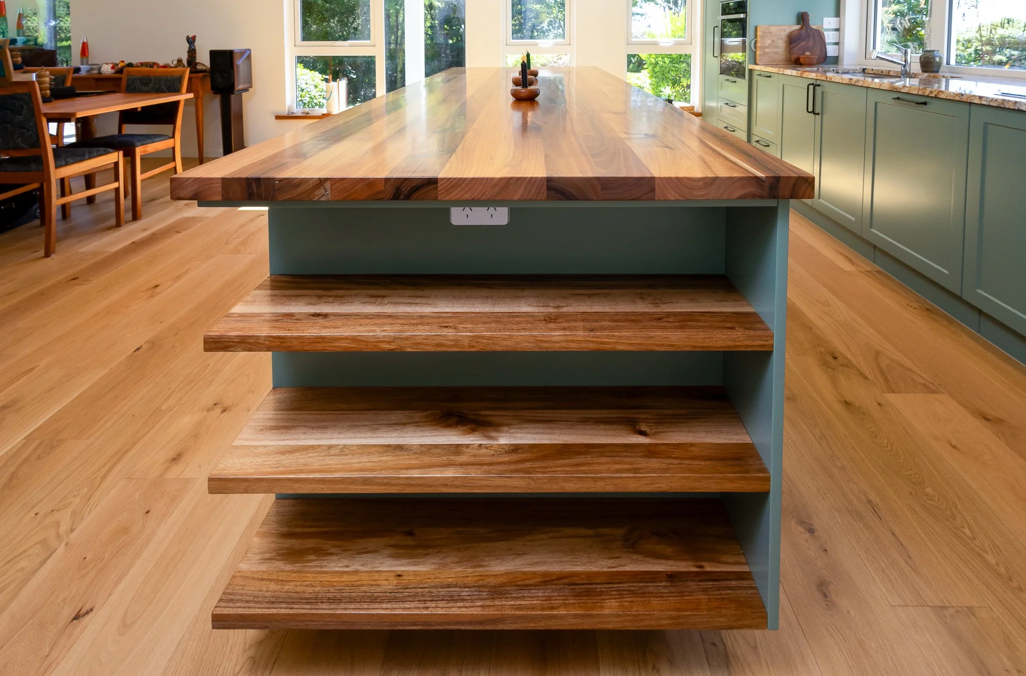 A modern wooden kitchen island with a natural finish, featuring a built-in electrical outlet on the side, in a bright kitchen with green cabinets and large windows.