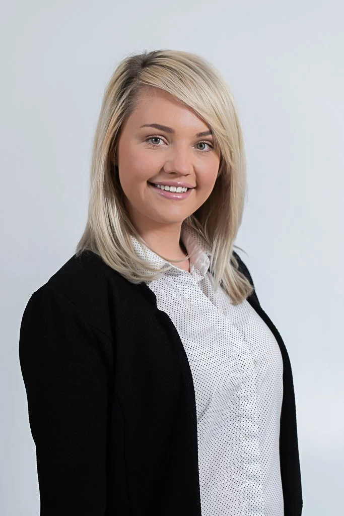 A young woman with blonde hair wearing a black blazer and a white collared shirt, smiling at the camera against a plain light gray background.